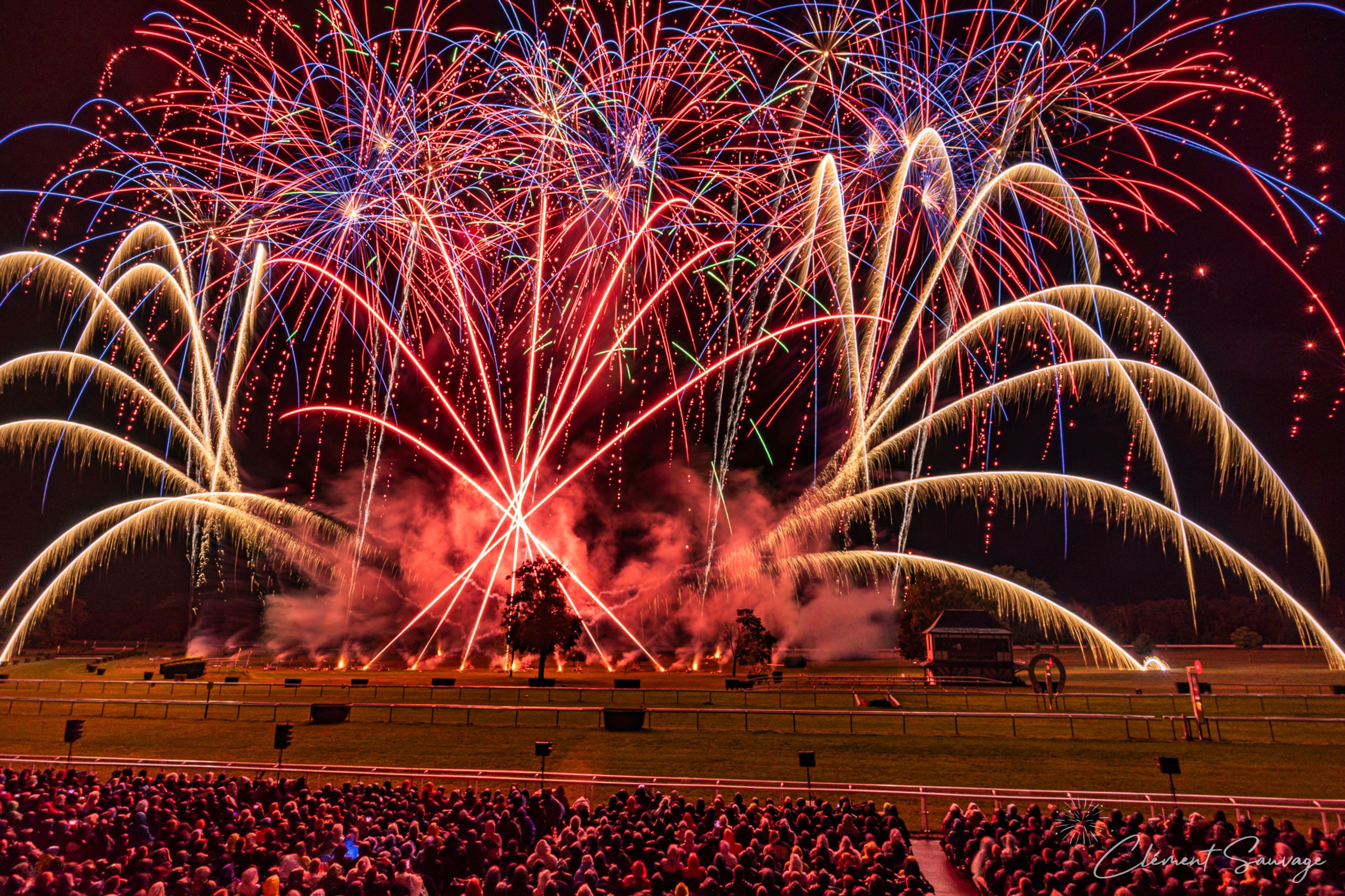 A large crowd watches a vibrant fireworks display at night during Masters de Feu 2025 in Compiègne, France, with red, blue, and golden fireworks illuminating the sky above a grassy field with scattered trees and a small structure.