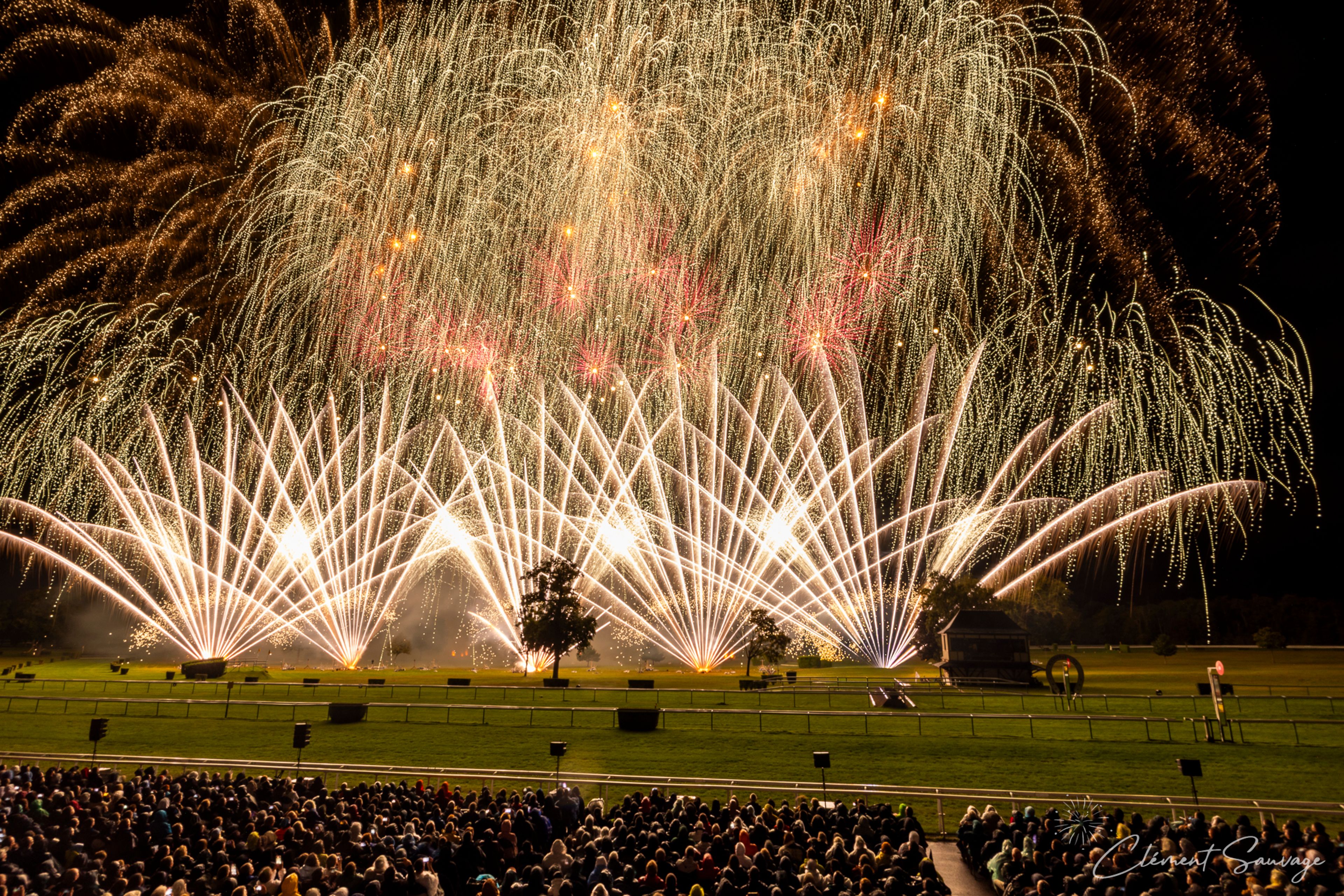 A large crowd watches a fireworks display at night during the Masters de Feu 2025 event in Compiègne, France, with bright golden and white fireworks creating fan-shaped and cascading patterns above a green field with a few trees and structures visible.