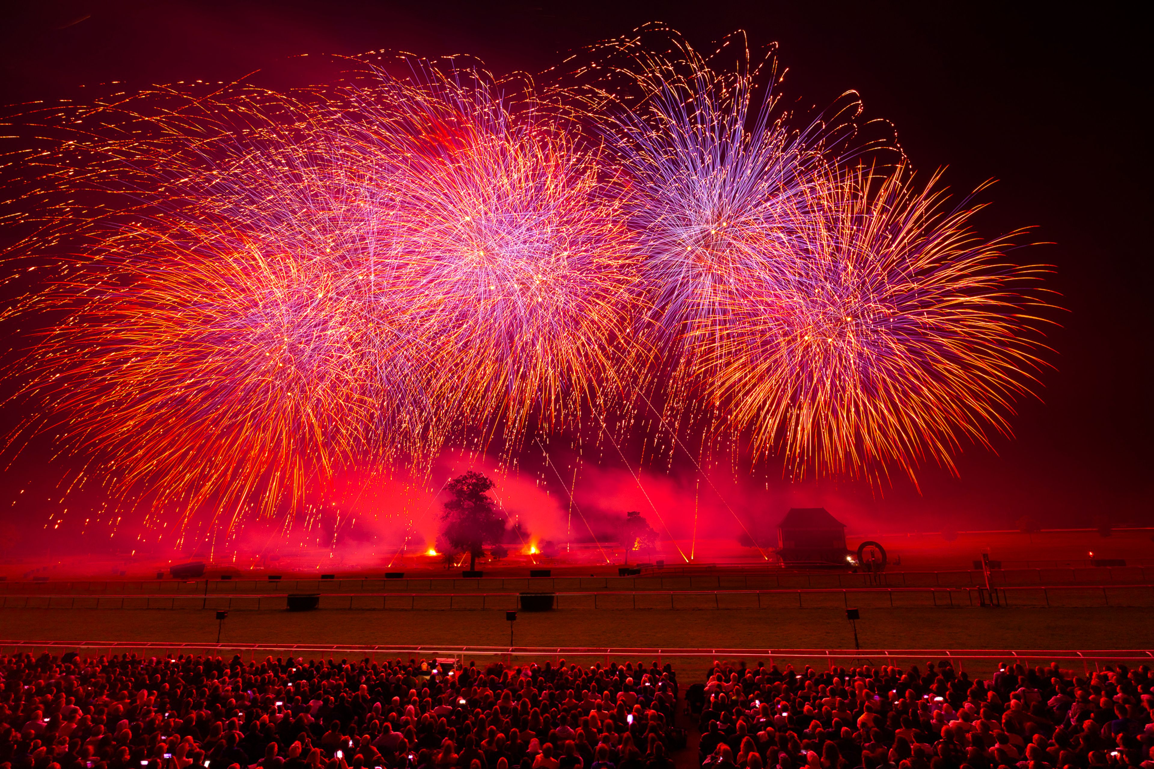 At the Masters de Feu 2024 event in Compiègne, France, a crowd watches a vibrant fireworks display with large bursts of red, orange, and purple light against a dark sky, illuminating smoke and silhouetted structures on the ground.