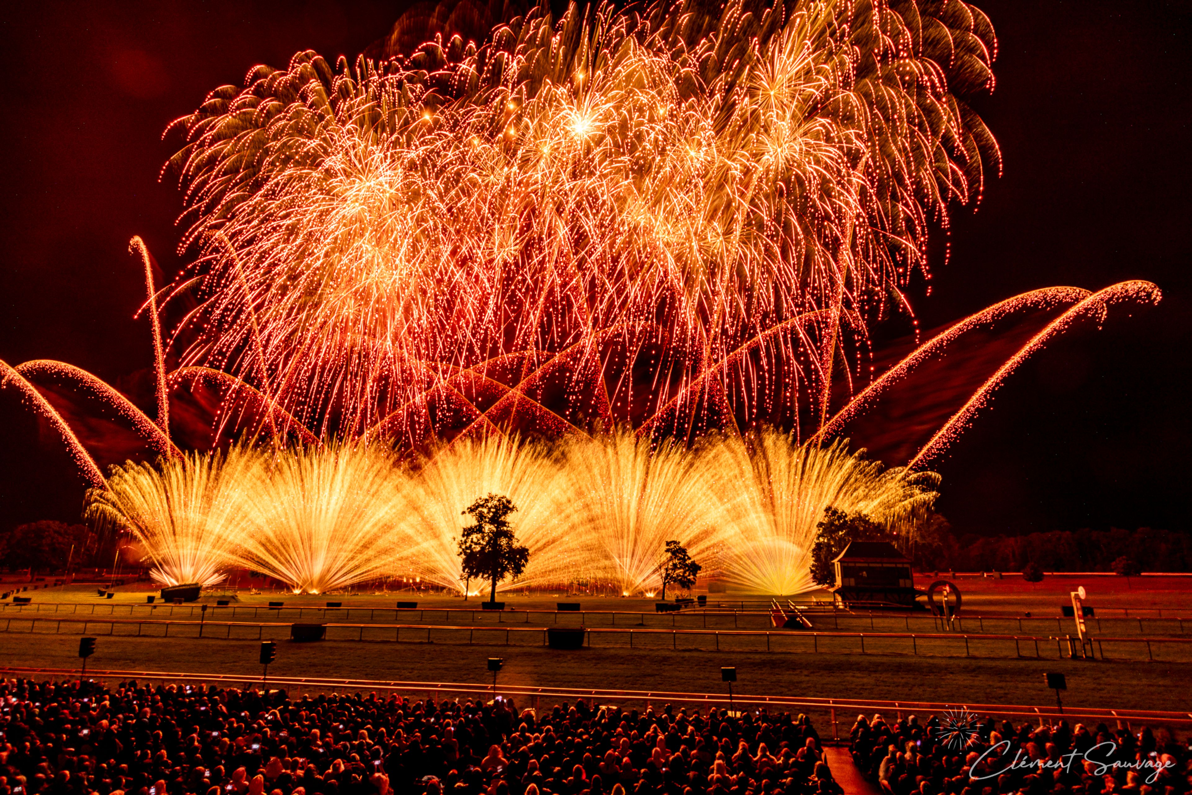 A large crowd watches a vibrant fireworks show at Masters de Feu 2025 in Compiègne, France, with bright orange and yellow bursts illuminating the night sky over an open field with silhouetted trees and structures.