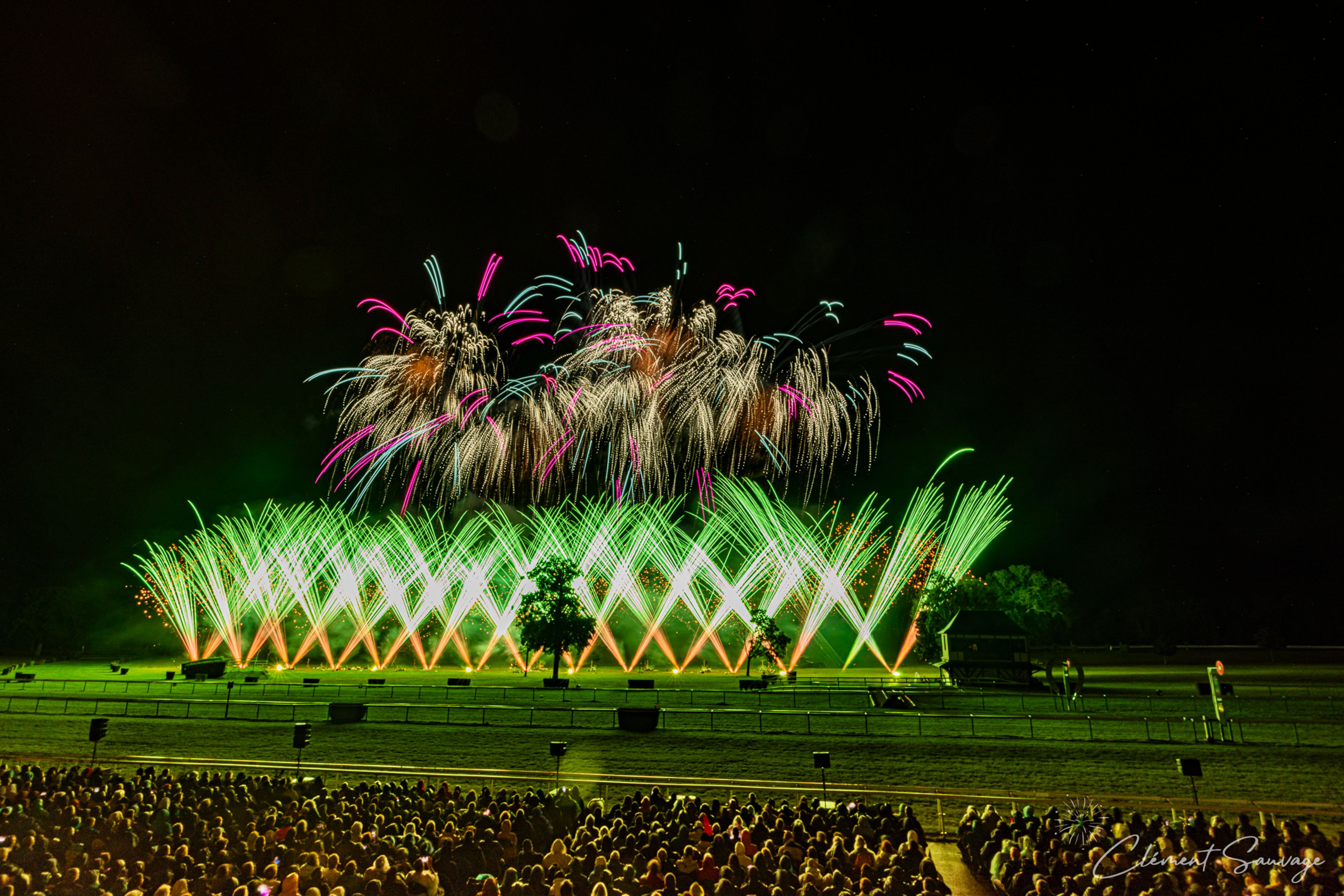 A night scene at Masters de Feu 2025 in Compiègne, France shows an audience watching a vibrant fireworks display featuring green, pink, white, and orange lights illuminating the sky and a field with trees and fence railings visible in the background.