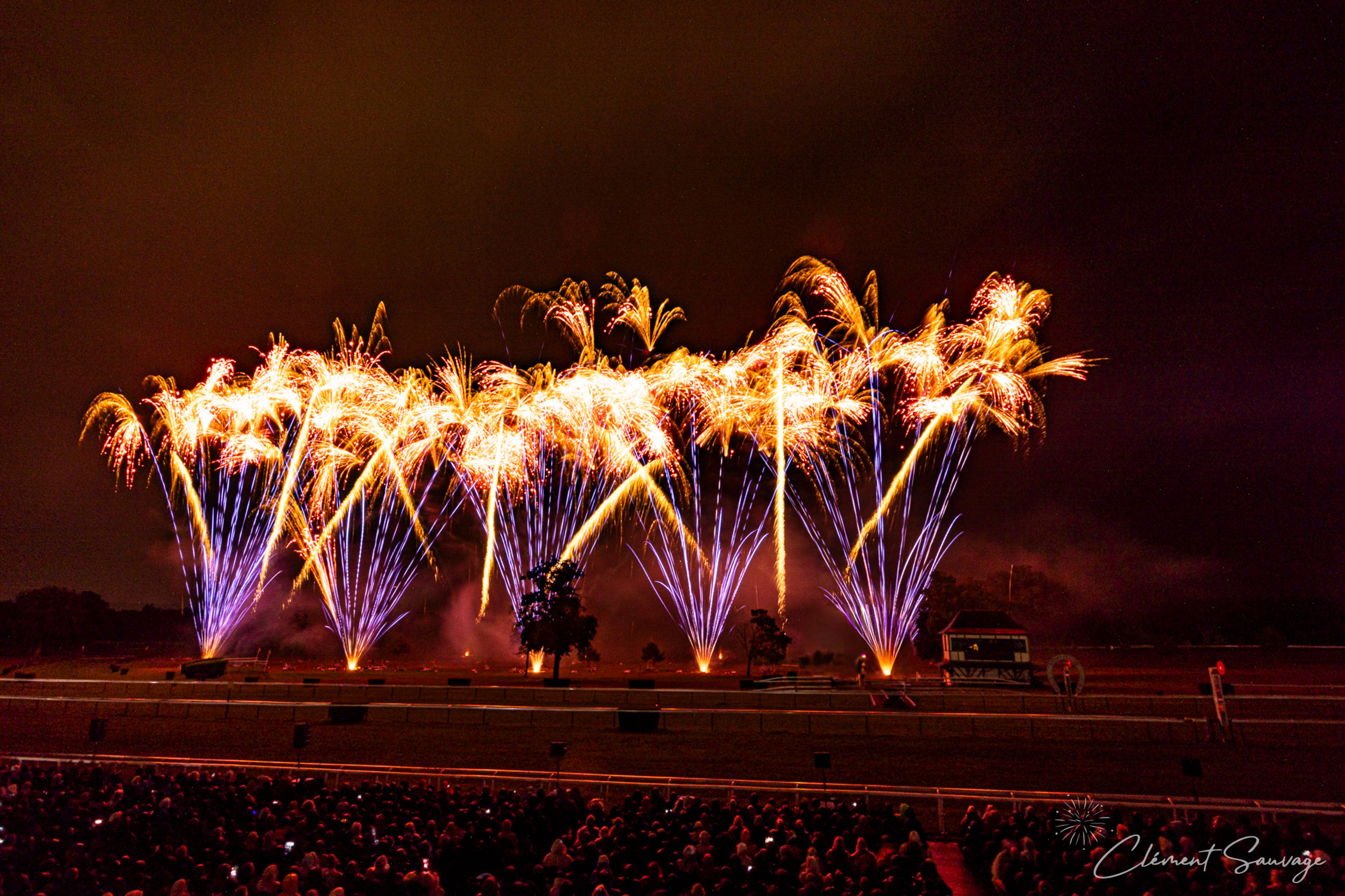 Bright orange and blue fireworks explode above a dark outdoor field with trees and a small structure, as a large audience watches from behind barriers during the Masters de Feu event on September 20, 2025, in Compiègne, France.