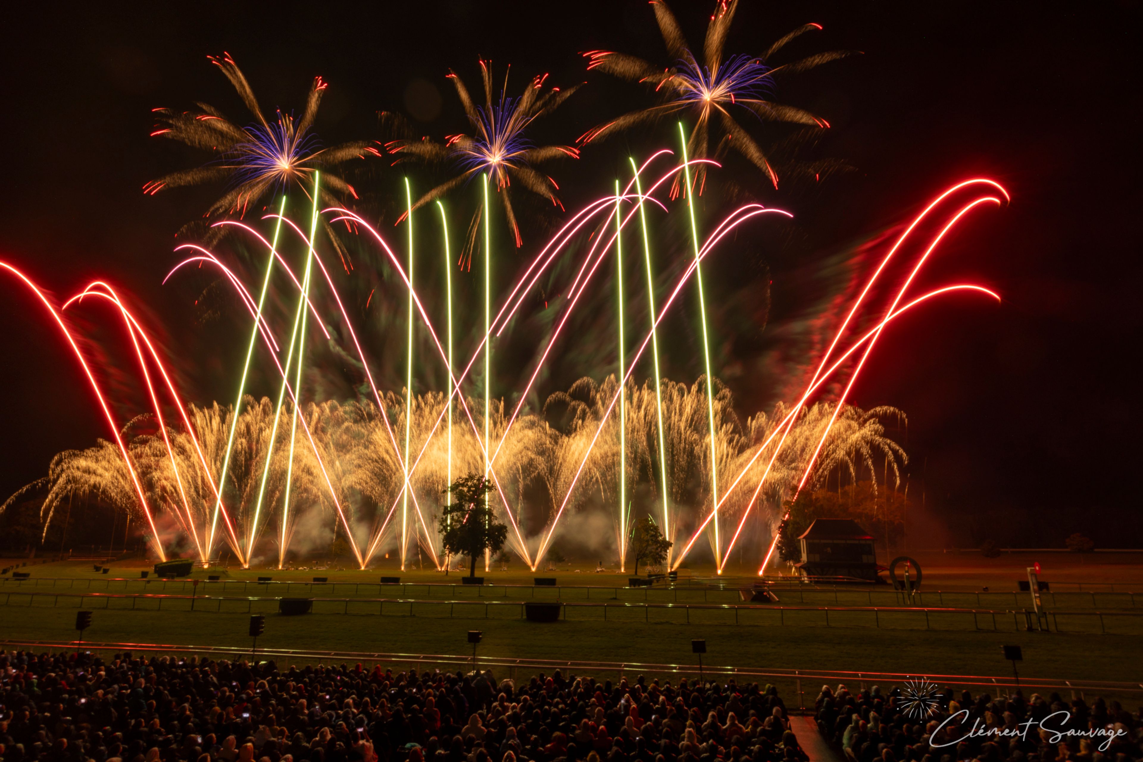 At the Masters de Feu event in Compiègne, France, spectators watch a nighttime fireworks display featuring red, green, blue, and gold bursting shapes and arcs above a grassy field with silhouetted trees, illuminated against the dark sky.