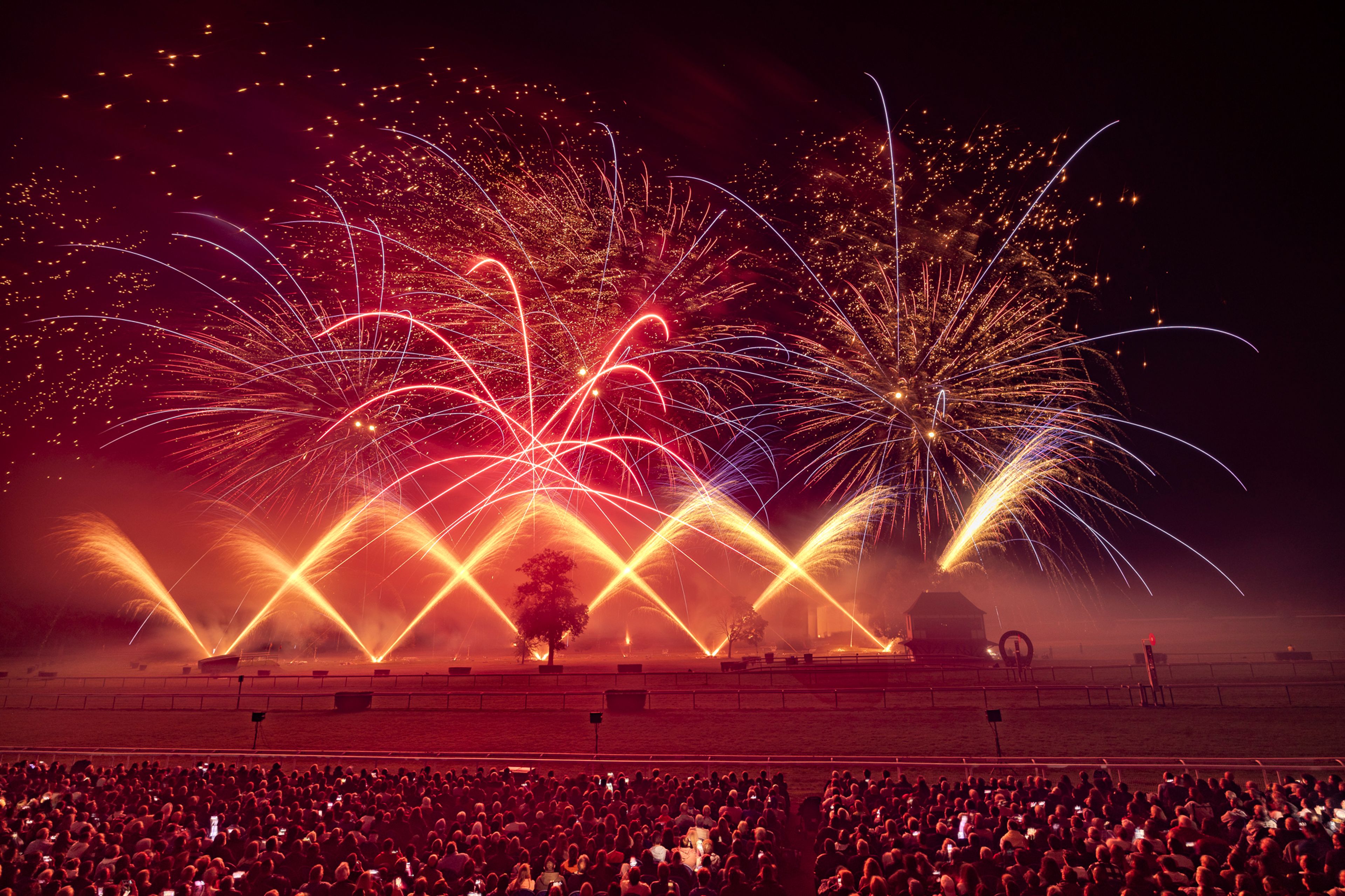 A large crowd watches a vibrant fireworks display with red, orange, yellow, and blue colors illuminating the night sky over an open field with some trees and small structures during the Masters de Feu event in Compiègne, France.