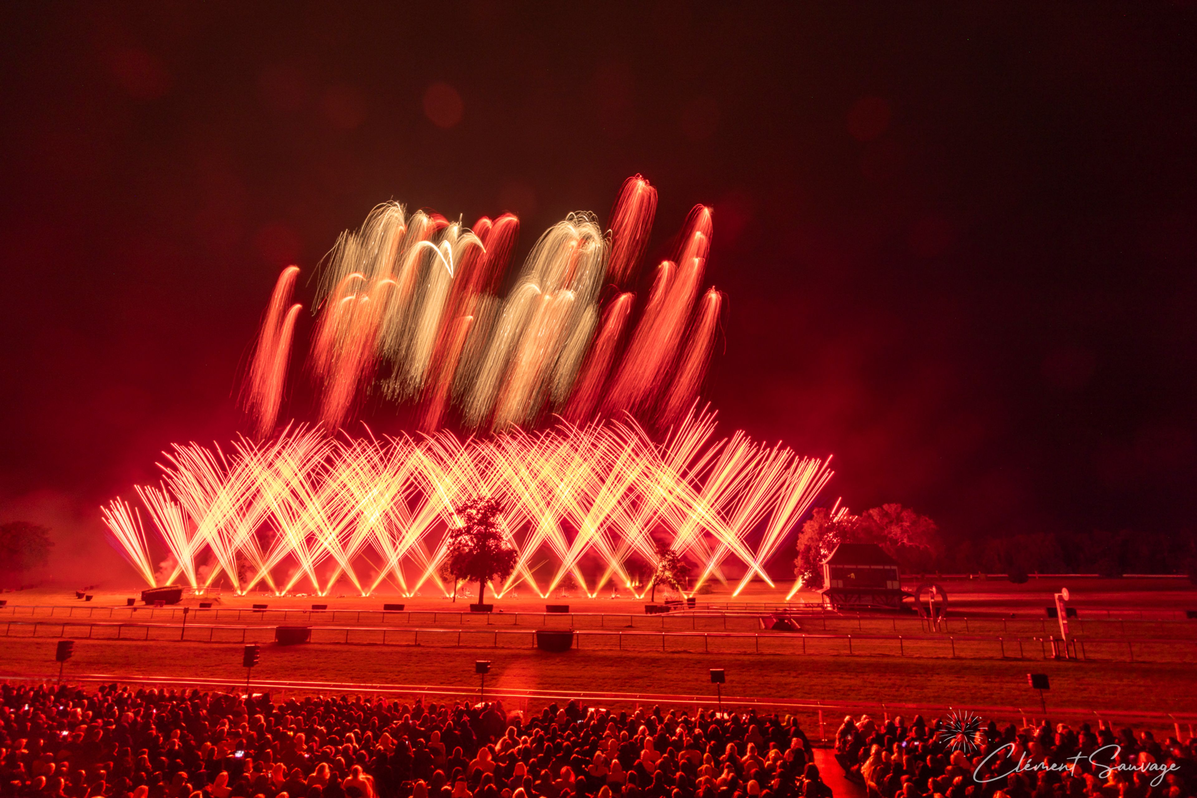 At Masters de Feu 2025 in Compiègne, France, a large crowd watches an intense fireworks display lighting up the night sky with vertical and arcing bursts of red and white light over an open field with some trees and structures visible in the background.