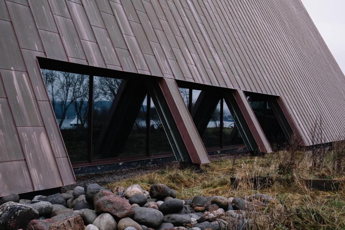 Foto av tak og fasade. Kobber taktekking møter glassfelt. Høstlandskap, vått gress og natursteiner. 