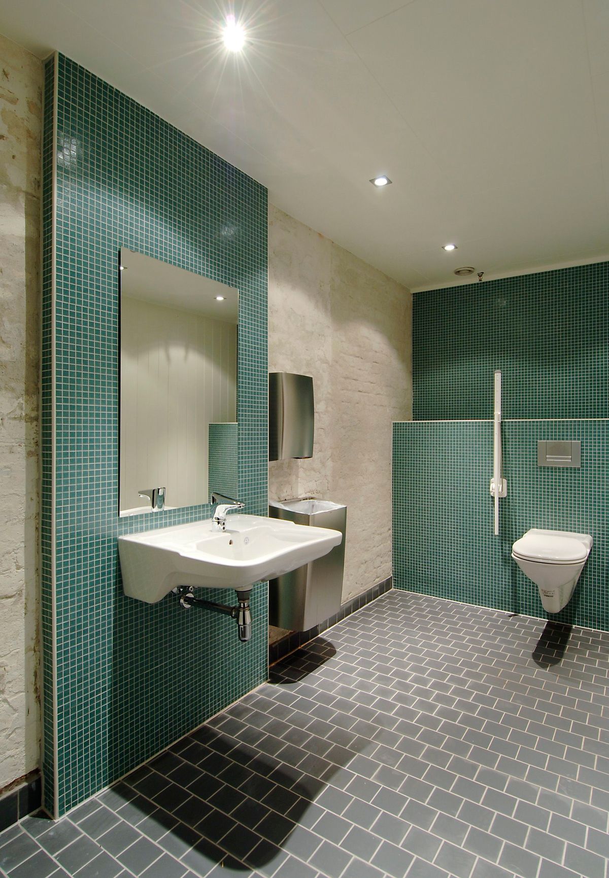 Foto av HC toalett, grønne fliser bak toalett og vask. Sort gulvfliser og hvitmalt tegl. 