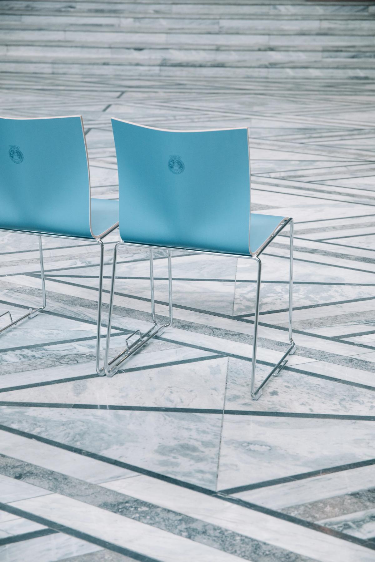Foto av 2 blå stoler. Stolene er av kryssfiner i en blå farge. Bein i polert metall med hengsler for stoler stilt i rekke. Oslo logo trykket i stolryggen. 