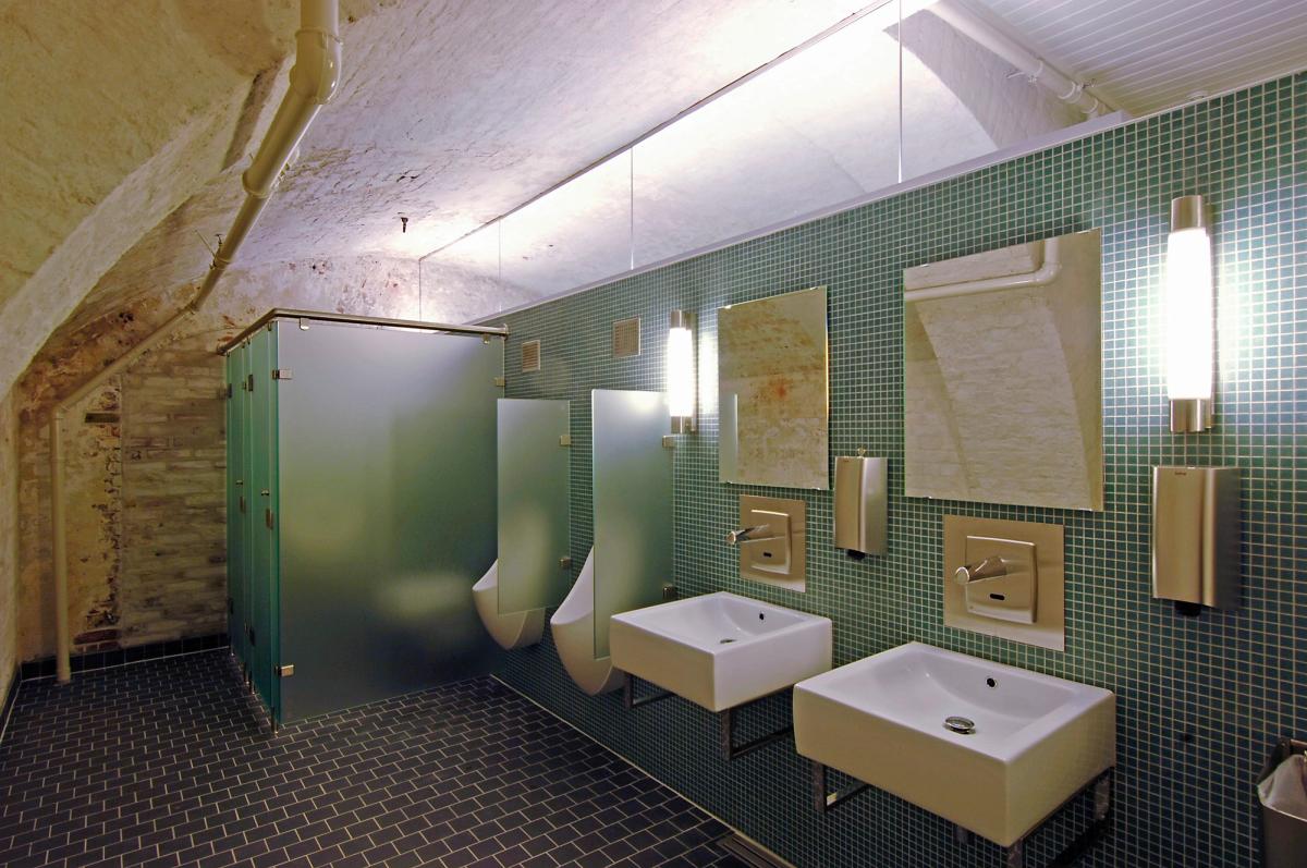 Foto av toalett situasjon i Hovedfortet, møtet mellom frostet glass, eksisterende hvit tegl, sorte gulvfliser og blå/grønn mosaikkfliser på wc og servant vegg. 