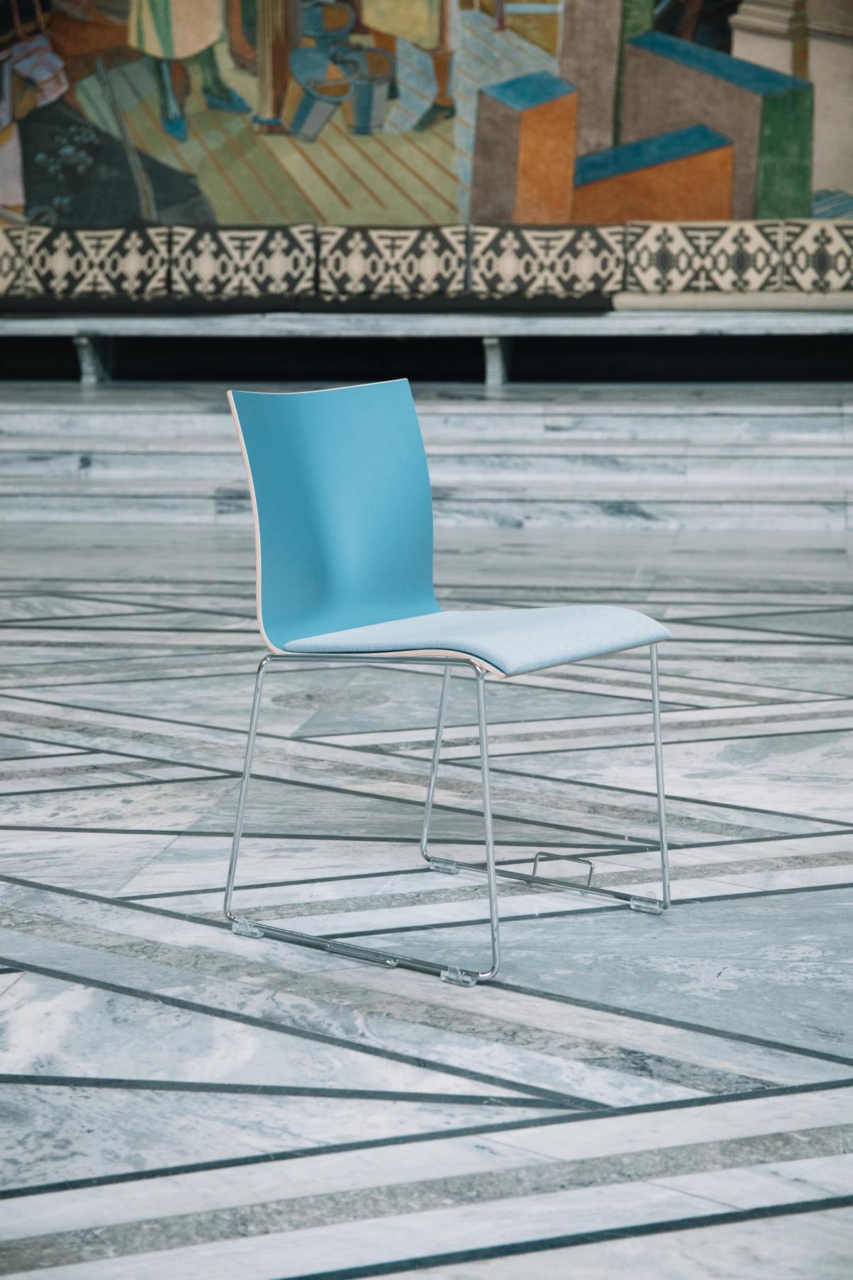 Foto av blå stol med tekstil trekk. Foto tatt på rådhuset. Stolen er av kryssfiner i en blå farge. Bein i polert metall med hengsler for stoler stilt i rekke. Blått tekstiltrekk som sete, avtagbart sete. 
