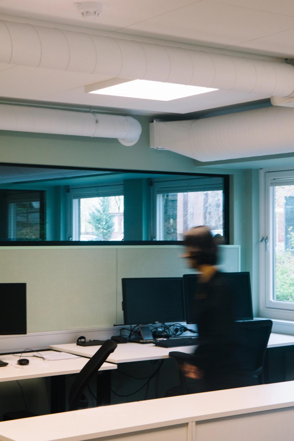 Foto av kontorlandskap med vindusrekke på høyre side, og glassfelt mellom kontorene for gjennomlys. Grønne vegger og veggabsorbenter. En person som går raskt forbi. 
