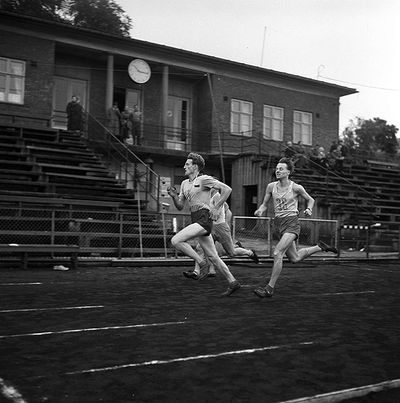 Historisk foto av idrettstevne. Tre løpere med klubbhuset i bakgrunnen.