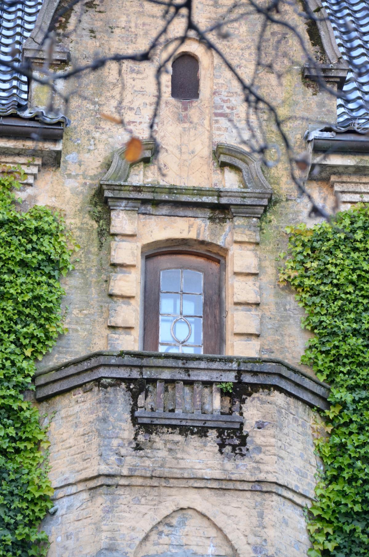 Foto av byggets fasade med fokus på balkong og vindu, omkranset av grønn vekst.