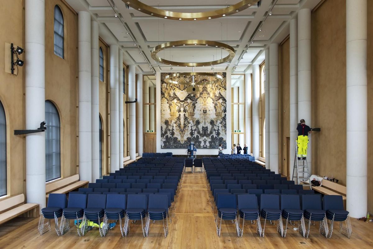 Foto ful bredde av aula. Blå stoler i ulike nyanser, gule vegger, hvitt tak og hvite søyler