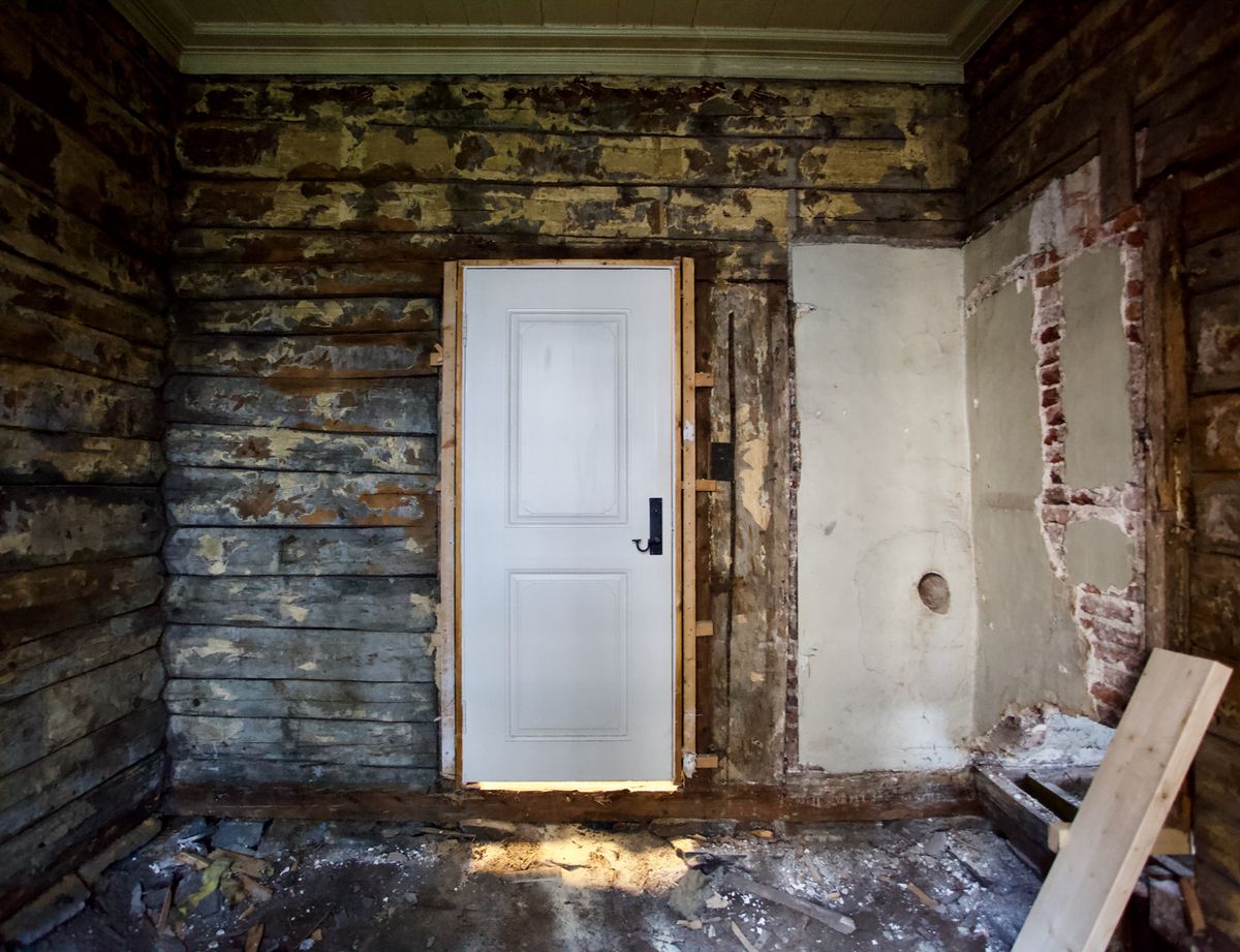 Foto av et rom før restaurering. Tømmervegger og murt tegl. Hvit dør uten karmer midt i bildet. 