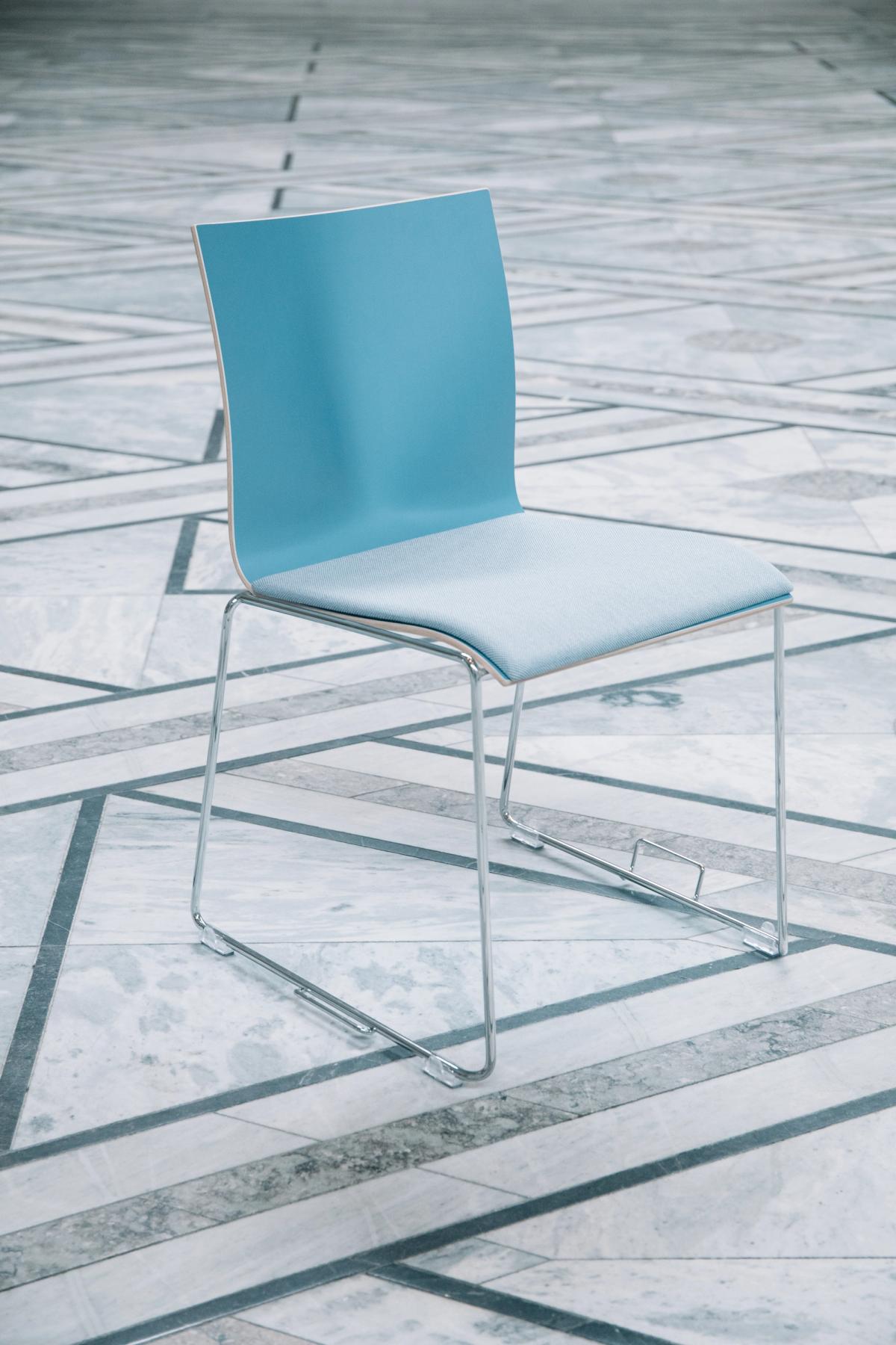 Foto av blå stol med tekstil trekk. Stolen er av kryssfiner i en blå farge. Bein i polert metall med hengsler for stoler stilt i rekke. Blått tekstiltrekk som sete, avtagbart sete. 