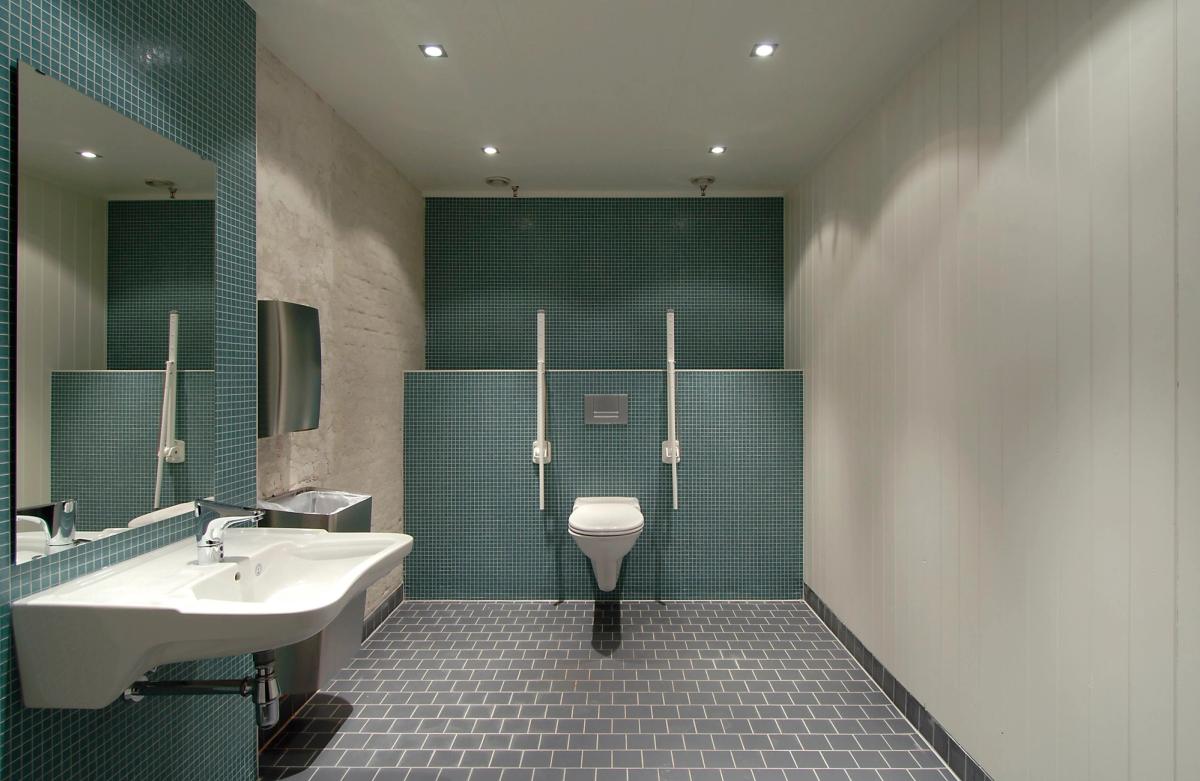 Foto av HC toalett, grønne fliser bak toalett og vask. Sort gulvfliser og hvitmalt tegl. 