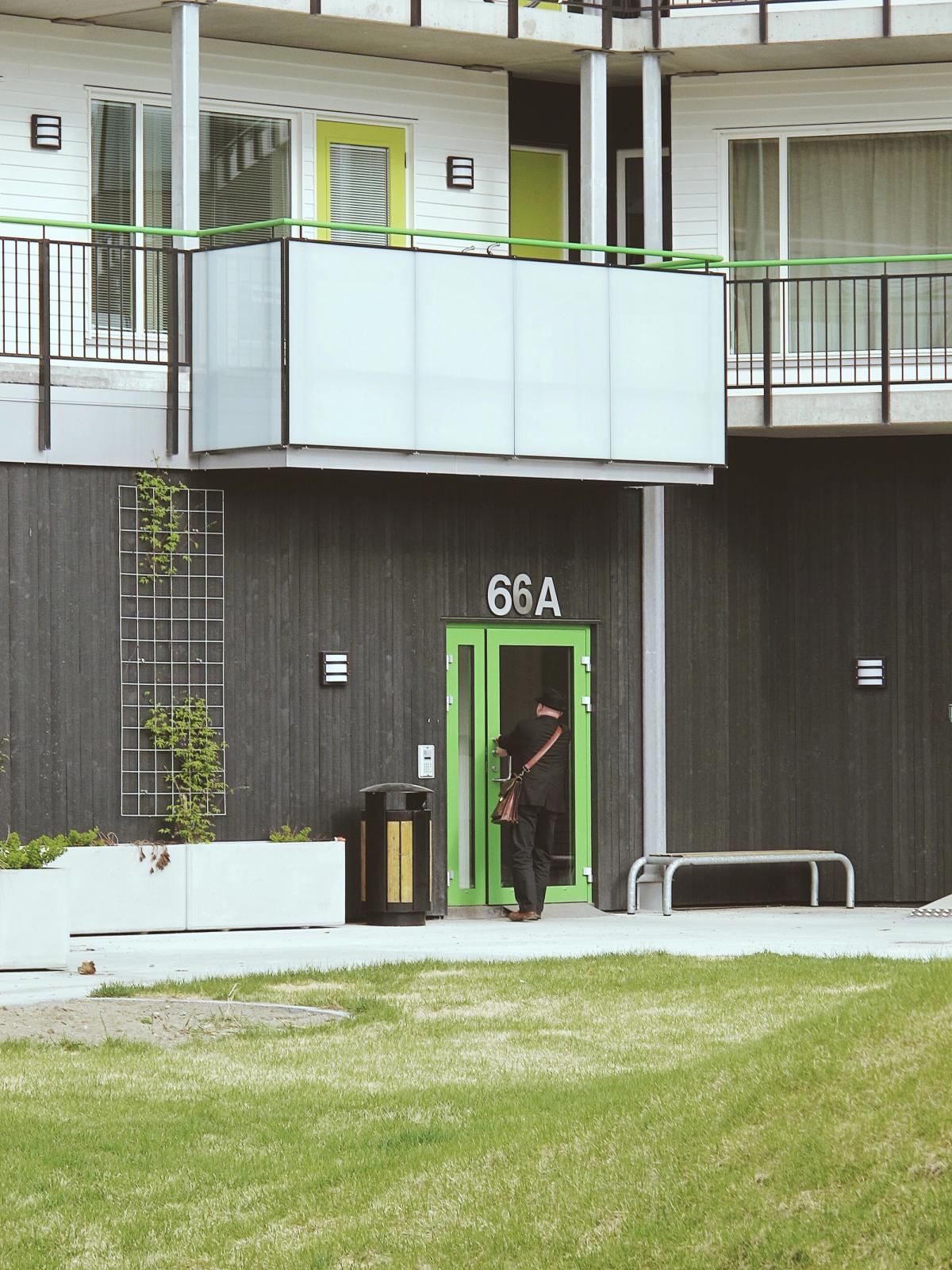 Foto av bakgården med grønn inngang og fasade i sort trekledning. Viser en person på vei inn døren.