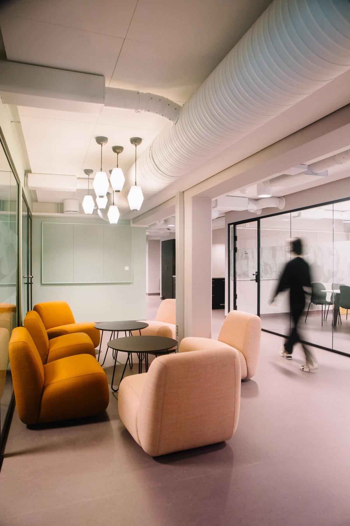 Foto av lunsjsone og kontorer i bakkant. Glassvegger eksponerer aktivitet i kontorene. Lilla gulv, grønne vegger, glassvegger og fargerike møbler.  Et menneske i bevegelse. 