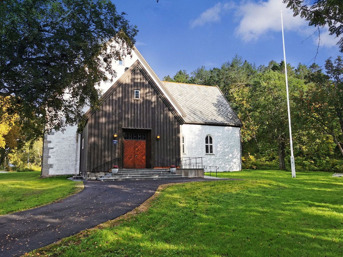 Foto av kirken på en fin sommerdag. Kirkens inngangsparti i sort trekledning og resterende vegger i hvitmalt stein. Rampe i skifer.