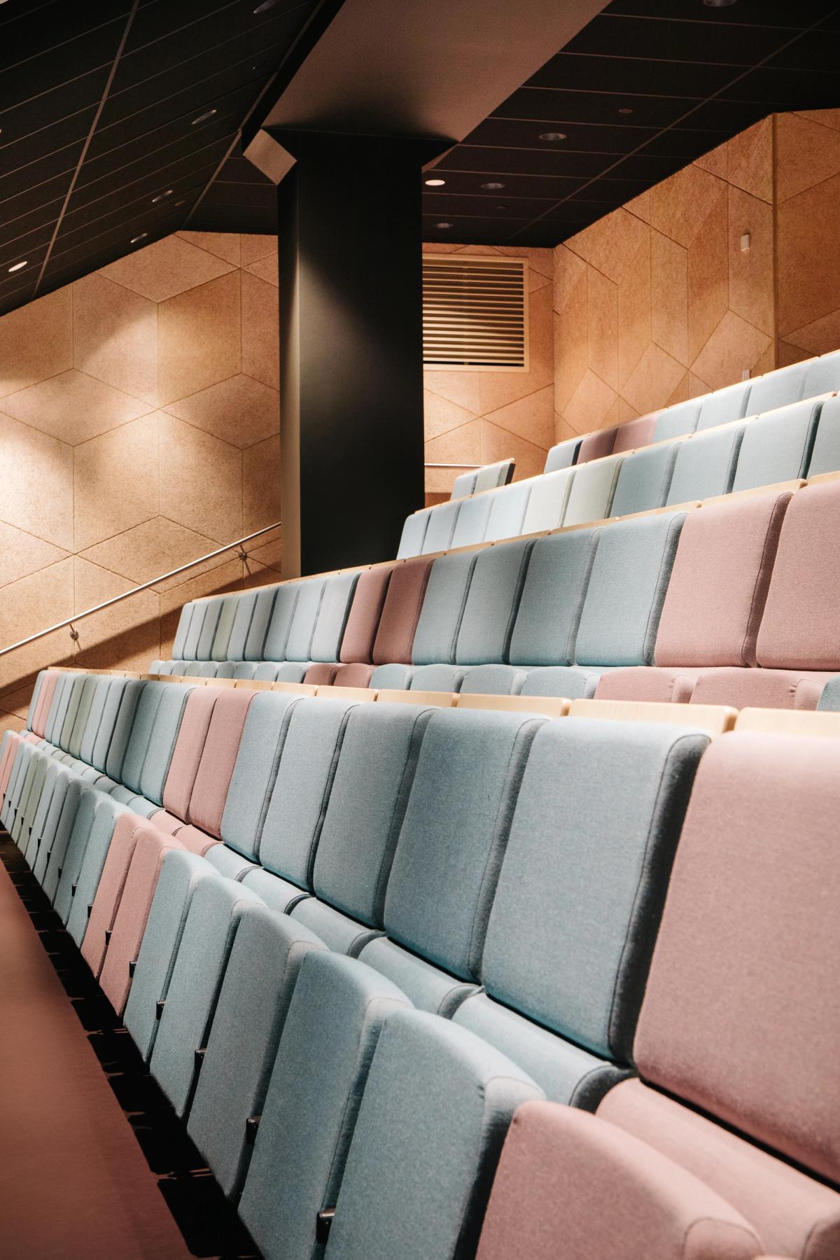 Foto av auditorium med stoler i rosa og blå, bakvegg i beig akustikkplater