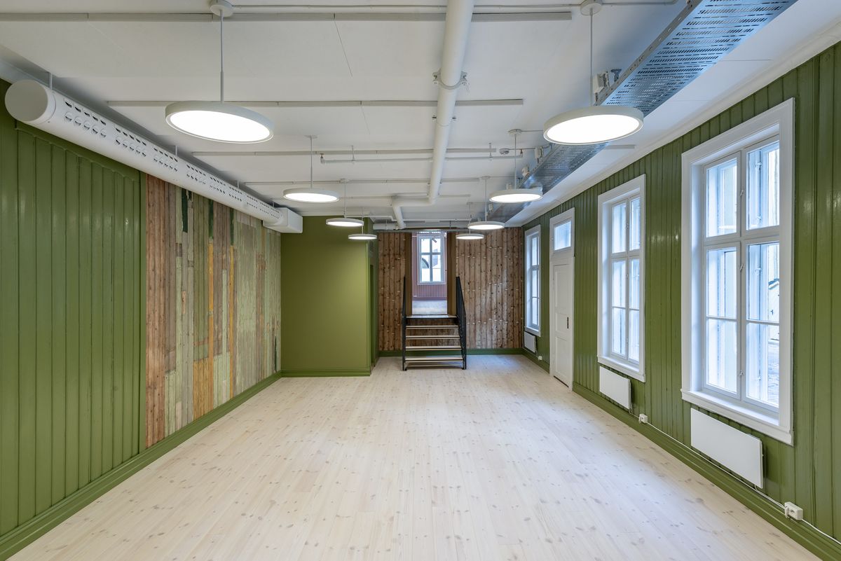 Fotografi av første større rom med grønne vegger og ubehandlet tre, med tilknyttning til et møterom i rosa. Trapp mellom rommene.
