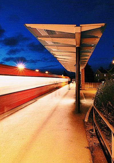Foto tatt på kveldstid av T-baneplattform, med t-bane i full fart. Mennesker i bevegelse på plattformen som er godt belyst. 