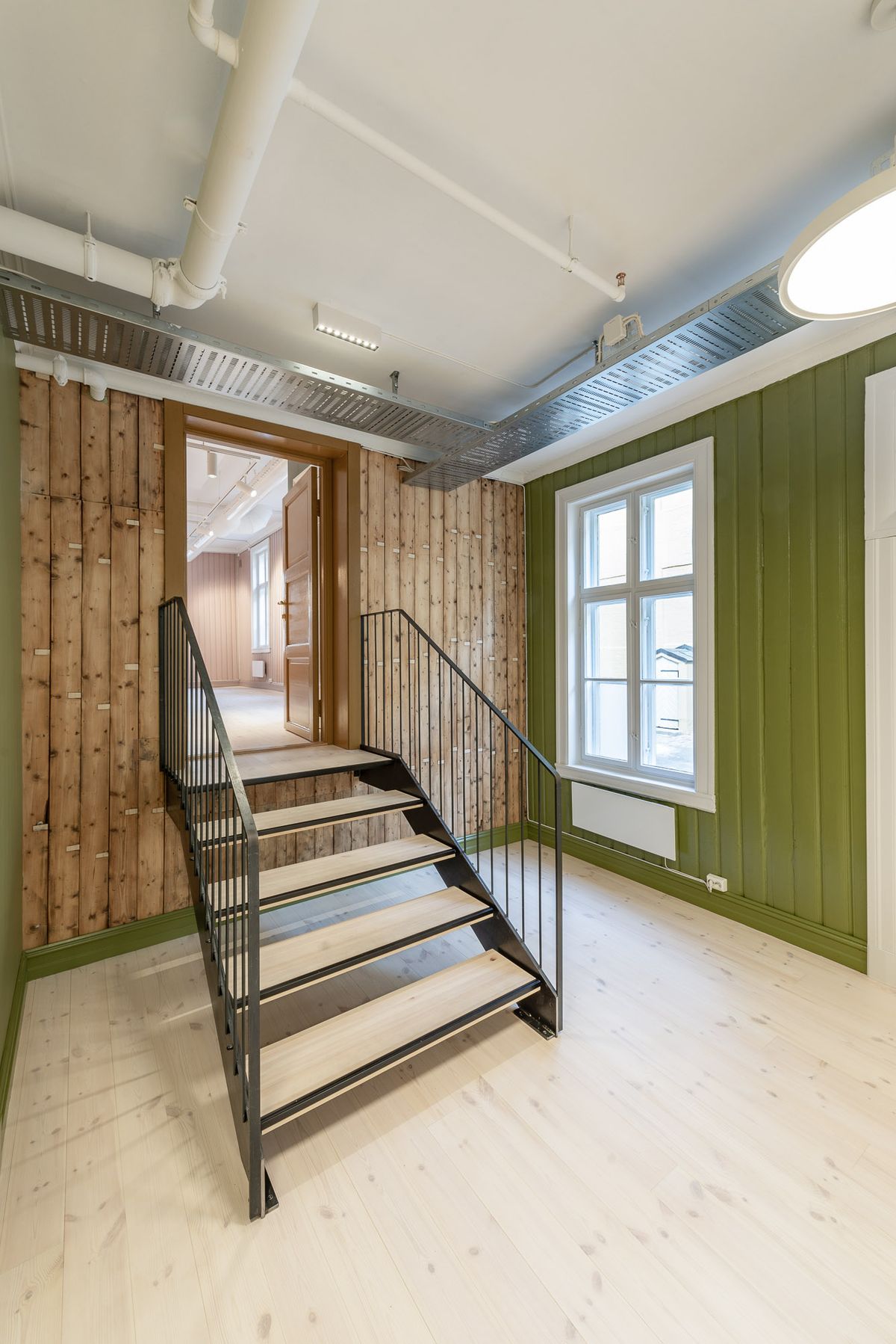 Fotografi av første større rom med grønne vegger og ubehandlet tre, med tilknyttning til et møterom i rosa. Trapp mellom rommene.