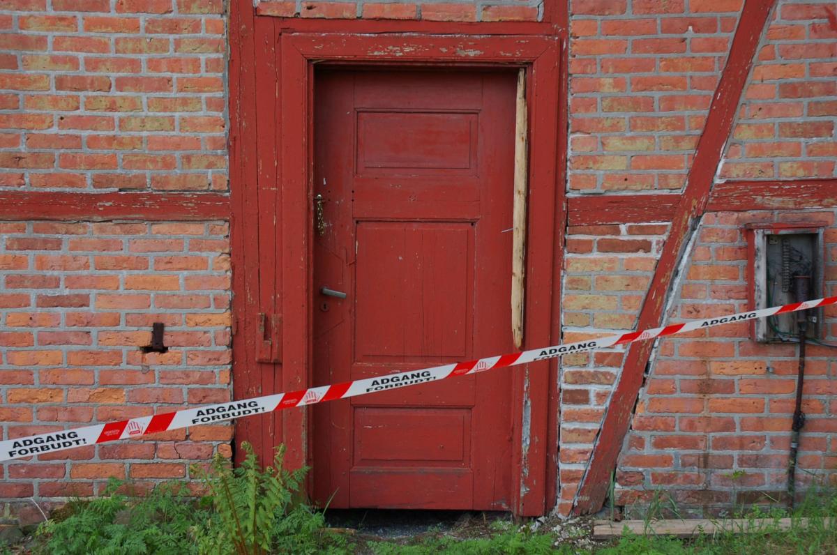 Foto av en falleferdig hus med "adgang forbud" bånd, rød dør i rød teglfasade.