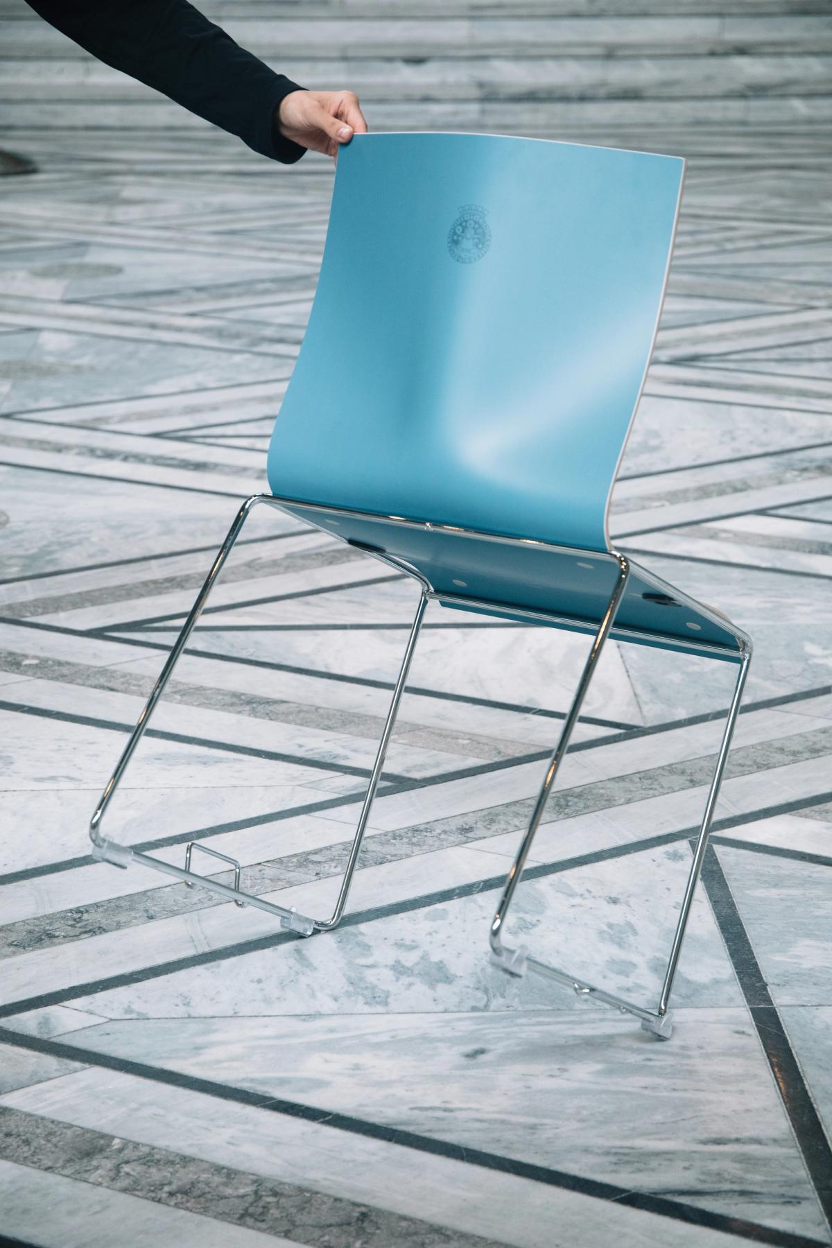Foto av blå stol på vippen. Stolen er av kryssfiner i en blå farge. Bein i polert metall med hengsler for stoler stilt i rekke. Et menneske vipper stolen for å vise undersiden. Oslo logo trykket i stolryggen. 