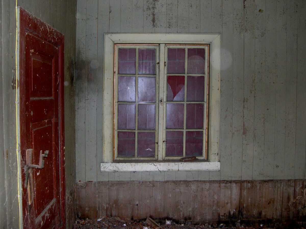 Foto av en interiør i falleferdig hus. Veggene er grønn panel, knust vindu og sliten rød dør.