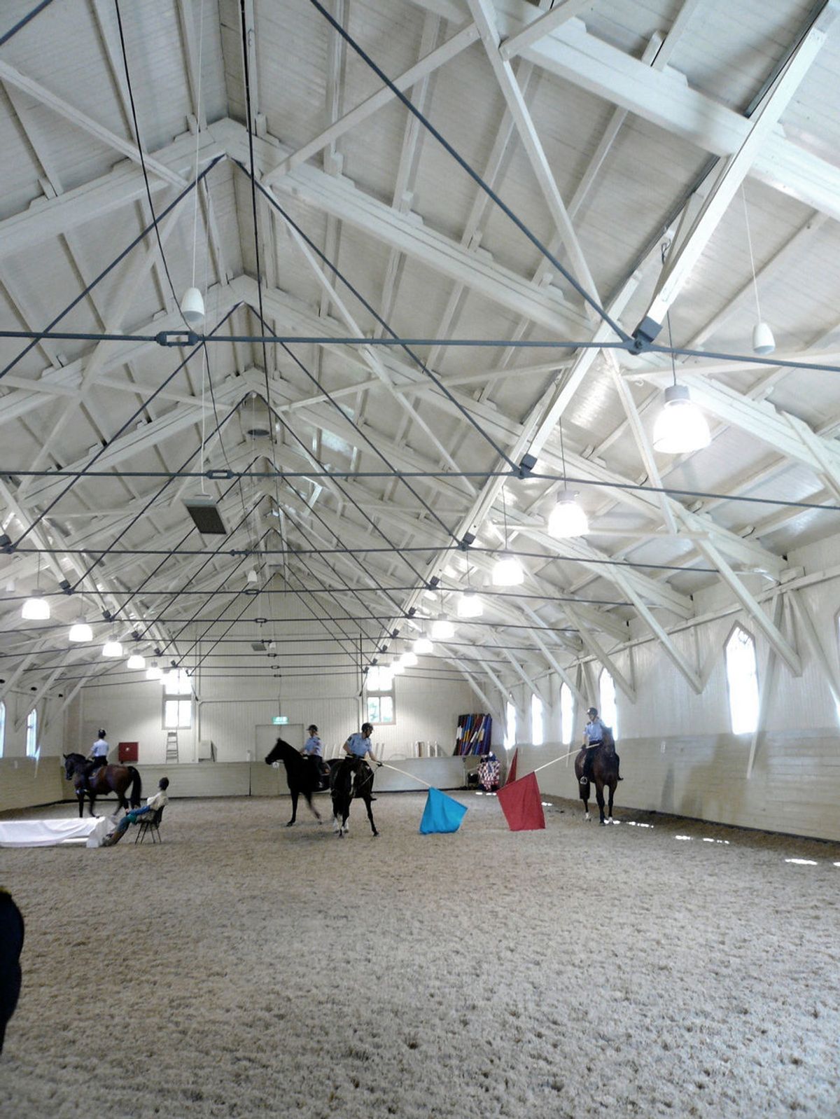 Foto av Rytterhusets interiør. Stor hall med hvite vegger og sand. Aktivitet i bilde med mennesker og hester. 