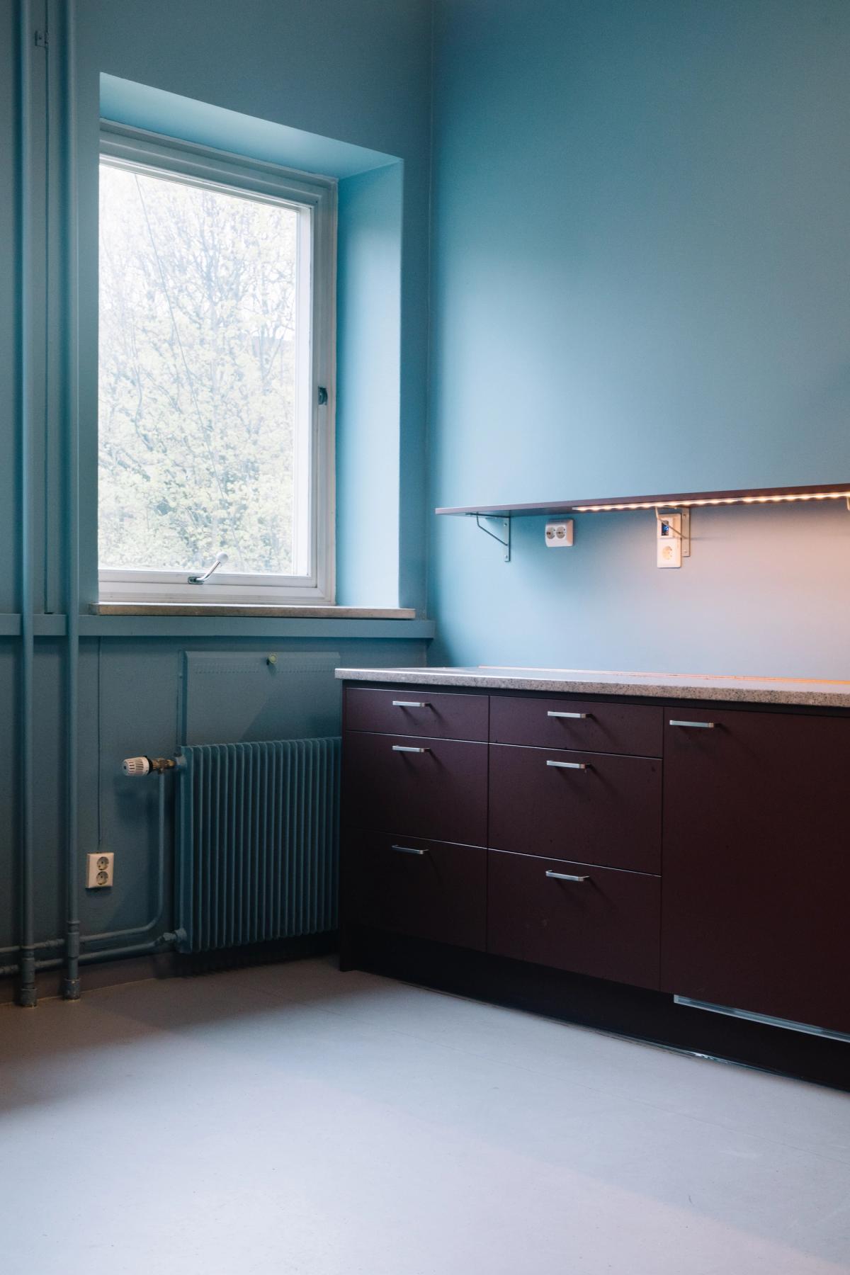 Foto av kjøkken for de ansatte i 2. etasje. Blå vegg og burgunder kjøkkenskap. Grorudstein som benkeplate, grå/rosa farge. 