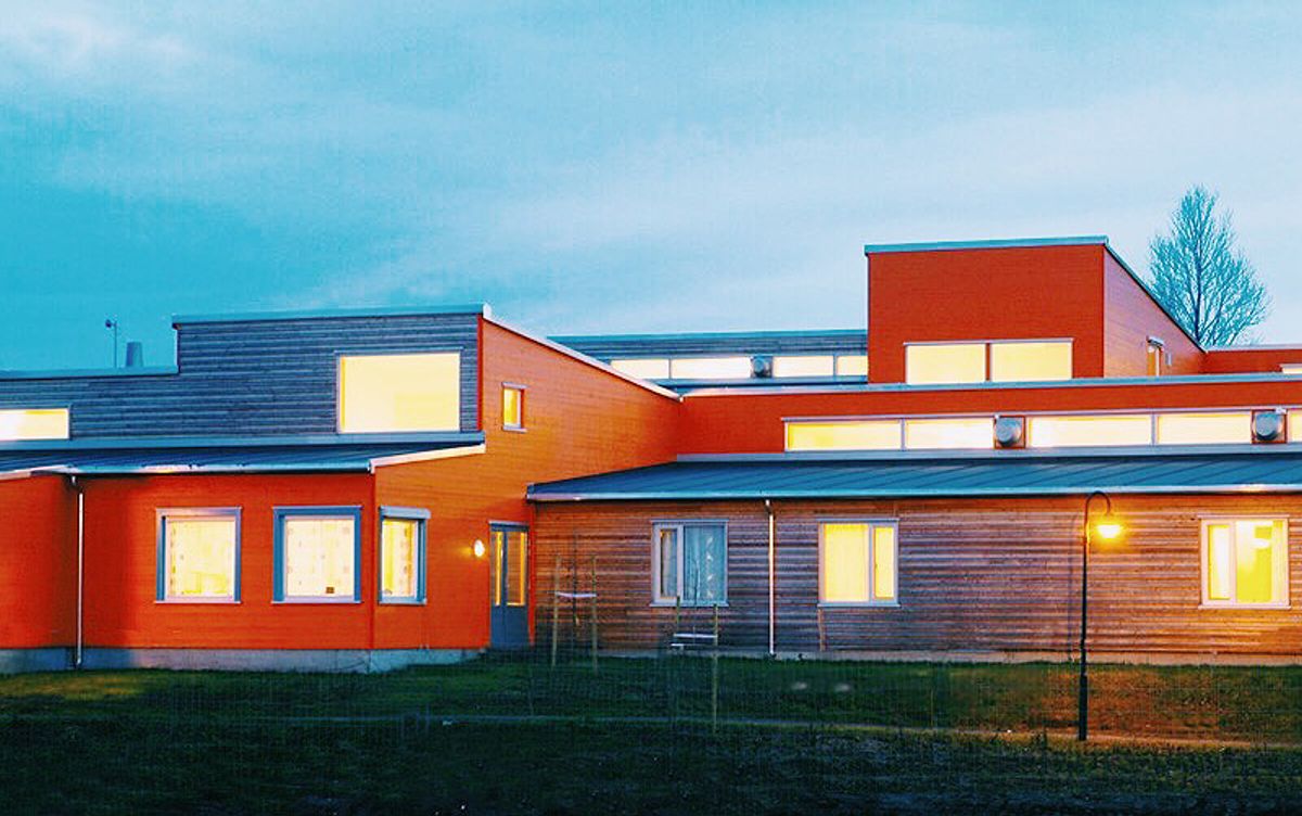 Foto av bygningsvolumer fra avstand med skrå tak. Fasader i orange treverk, med vinduer i ulike formater. 