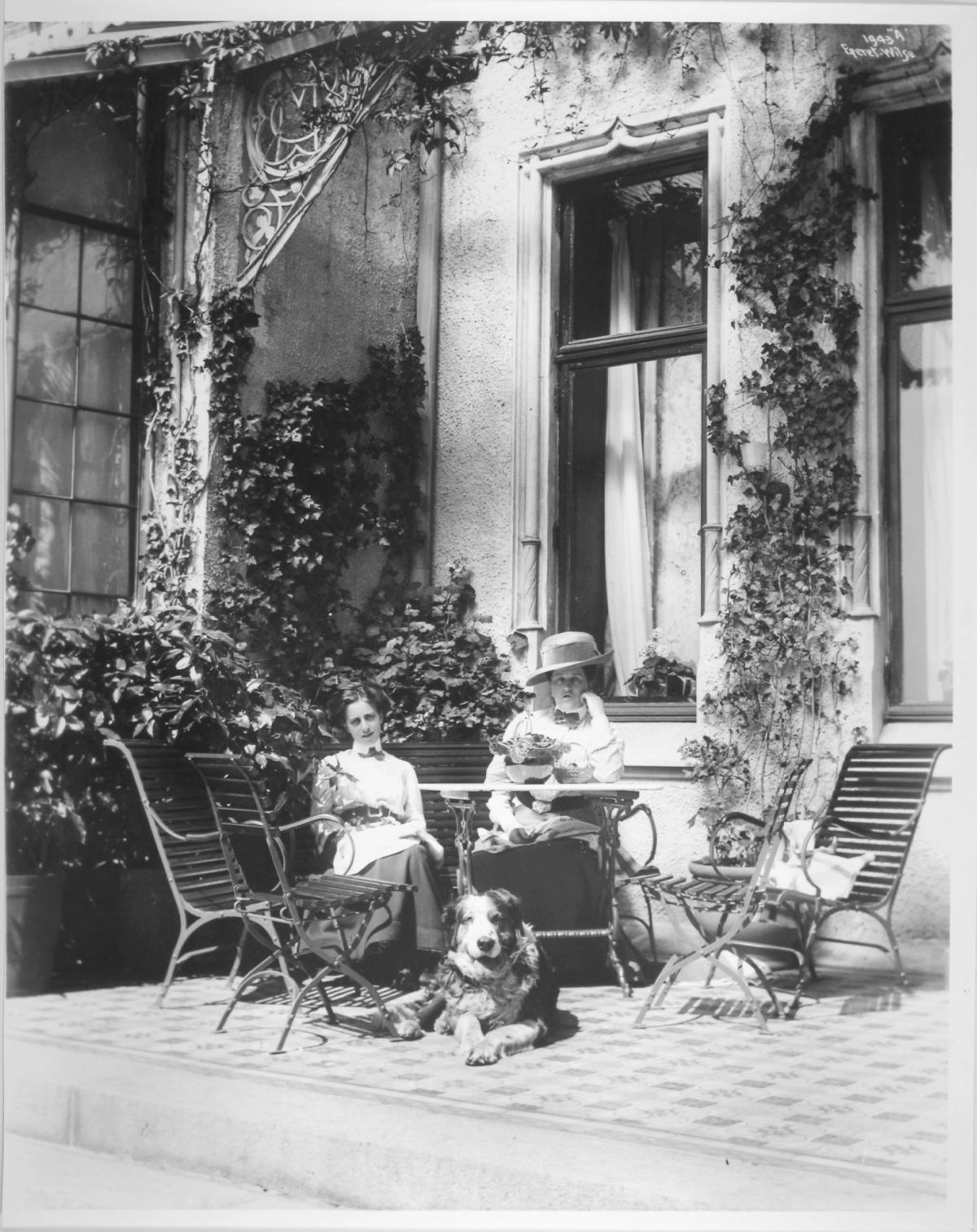 Historisk foto av Gamlehaugen. Fotografiet viser huset i bakgrunn av to damer på terrassen sammen med en hund. WB Nilsen 1910. 