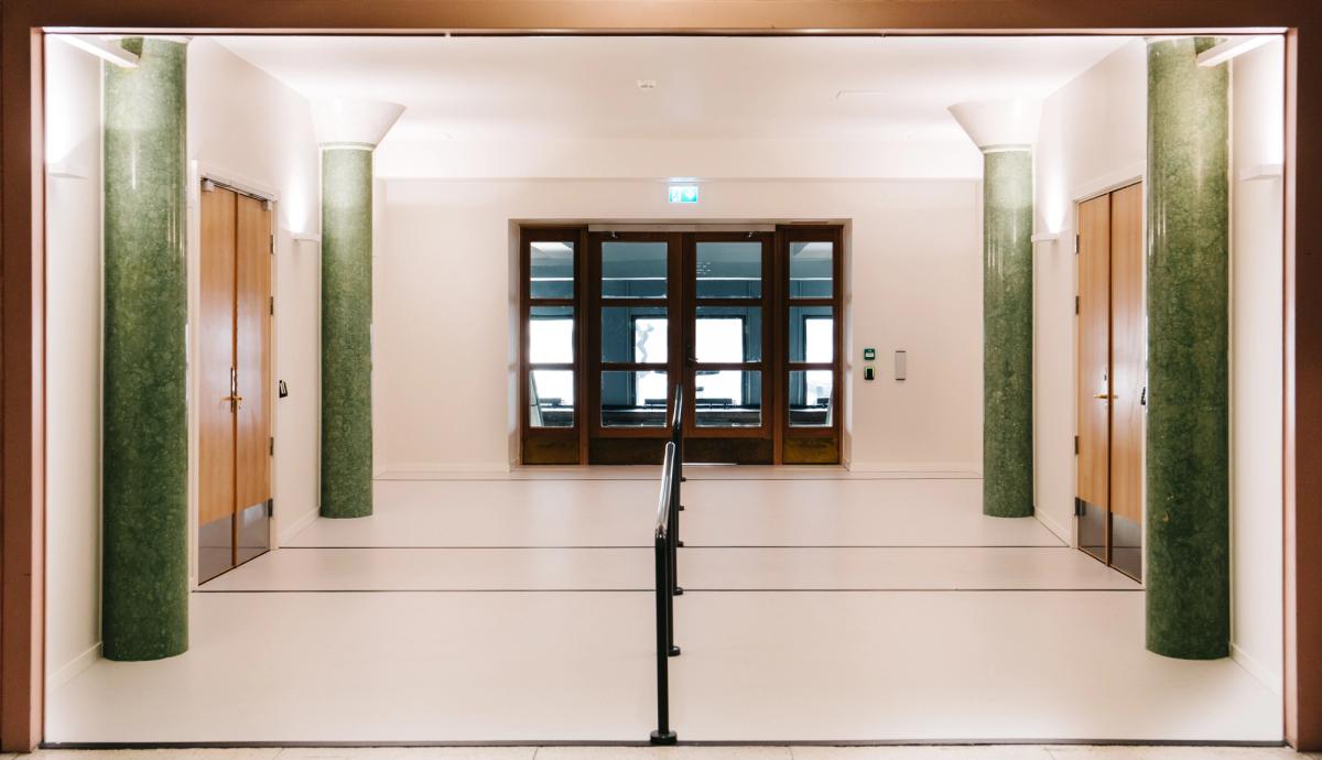 Foto av korridor fra Oslo Rådhus hallen til møteromssenter. Hvite vegger, hvitt gulv og grønne søyler. 