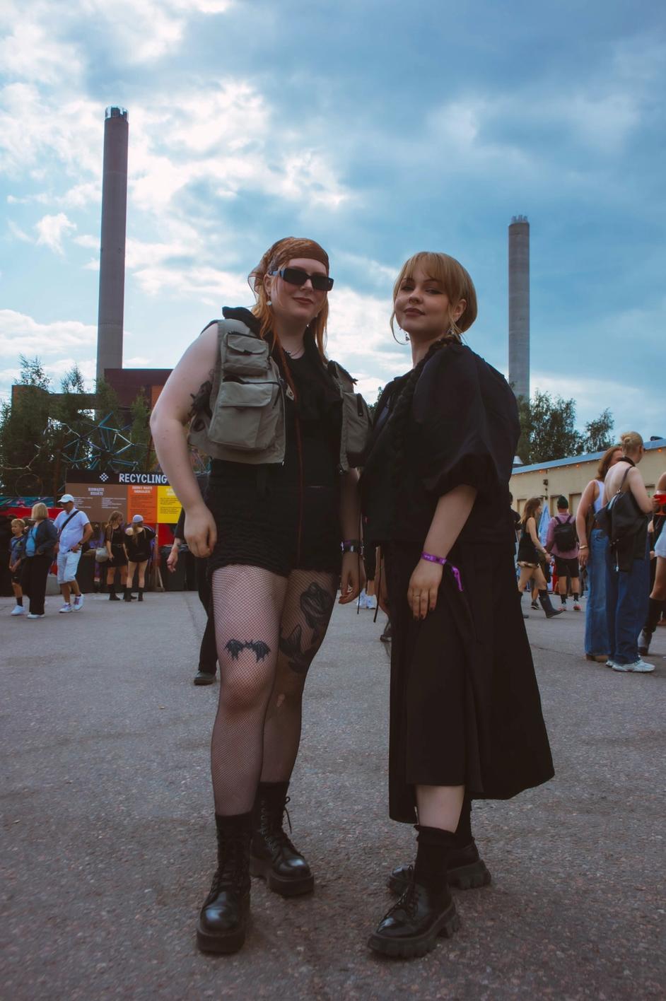 Две девушки на фестивале в тёмной одежде на фоне индустриальных труб (Kaksi naista festivaalilla tummissa vaatteissa, taustalla piiput).