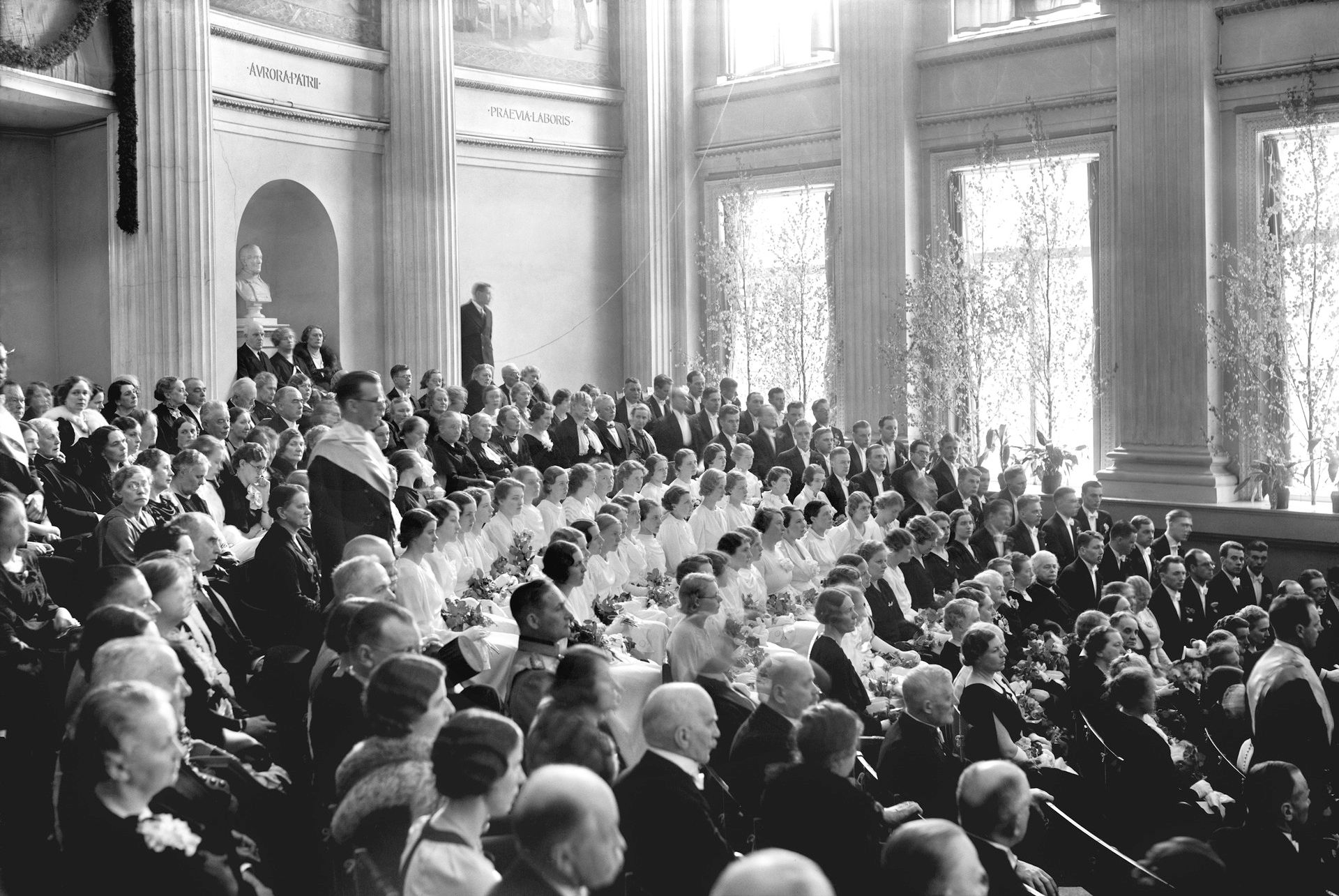 Mustavalkoinen valokuva Helsingin yliopiston promootiosta vuonna 1939 juhlasalissa. Salissa istuu suuri joukko juhlavasti pukeutuneita ihmisiä, keskellä nuoria valkoisiin pukeutuneita promovoitavia, taustalla juhlapukuista yleisöä. Tilaa koristavat korkeat ikkunat, pylväät ja keväiset koivunoksat.