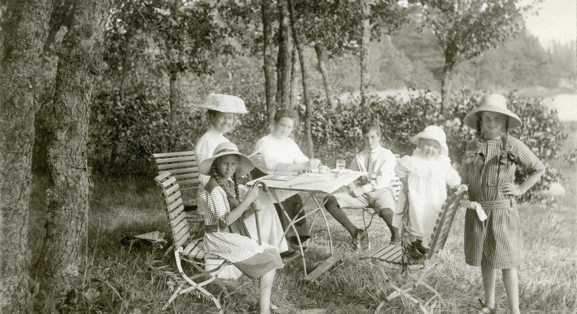 Valokuva 1900-luvulta näyttää kuusi henkilöä, aikuisia ja lapsia viettämässä kesäpäivää ulkona puiden katveessa. Osalla on yllään 1900-luvun alun tyyliset vaatteet ja leveälieriset hatut. Taustalla näkyy metsää.