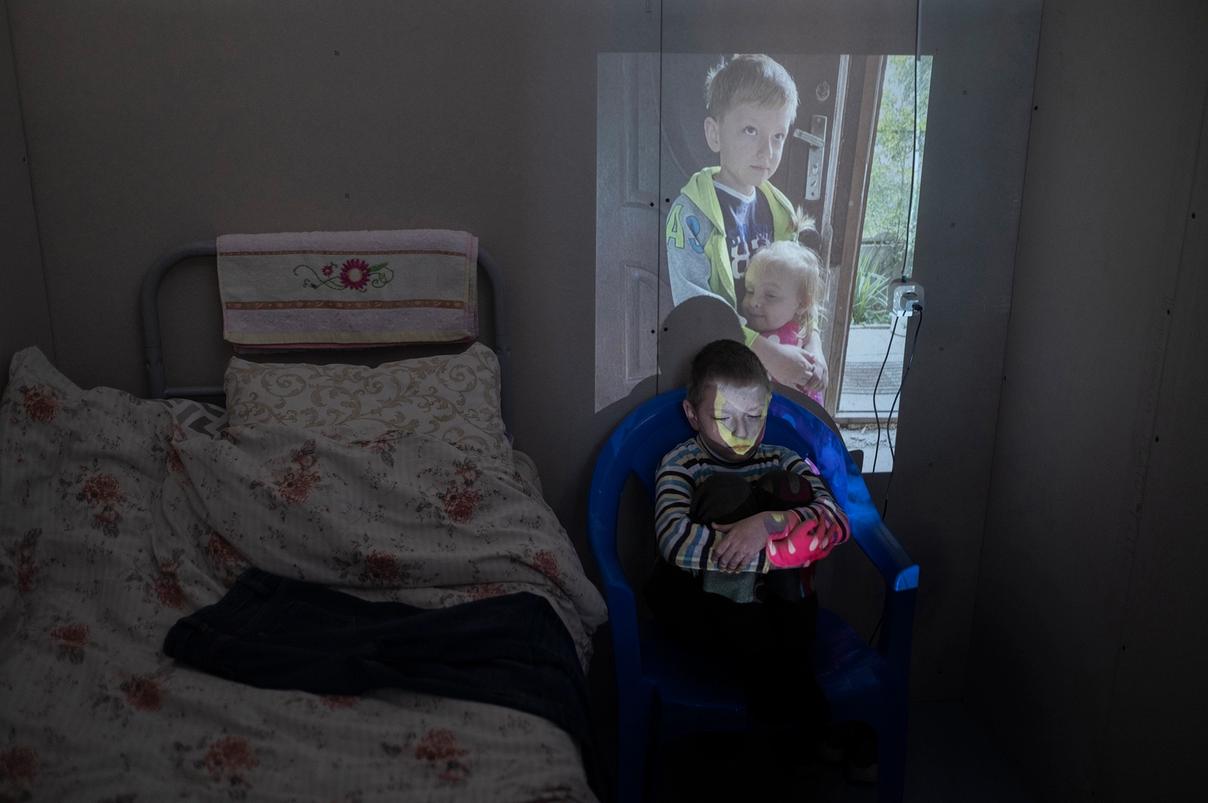 Мальчик сидит в кресле, обняв колени, на его лицо проецируется фотография детей, обнимающихся у двери (Poika istuu tuolissa polviaan halaillen, hänen kasvoilleen heijastuu kuva halaavista lapsista oven luona)