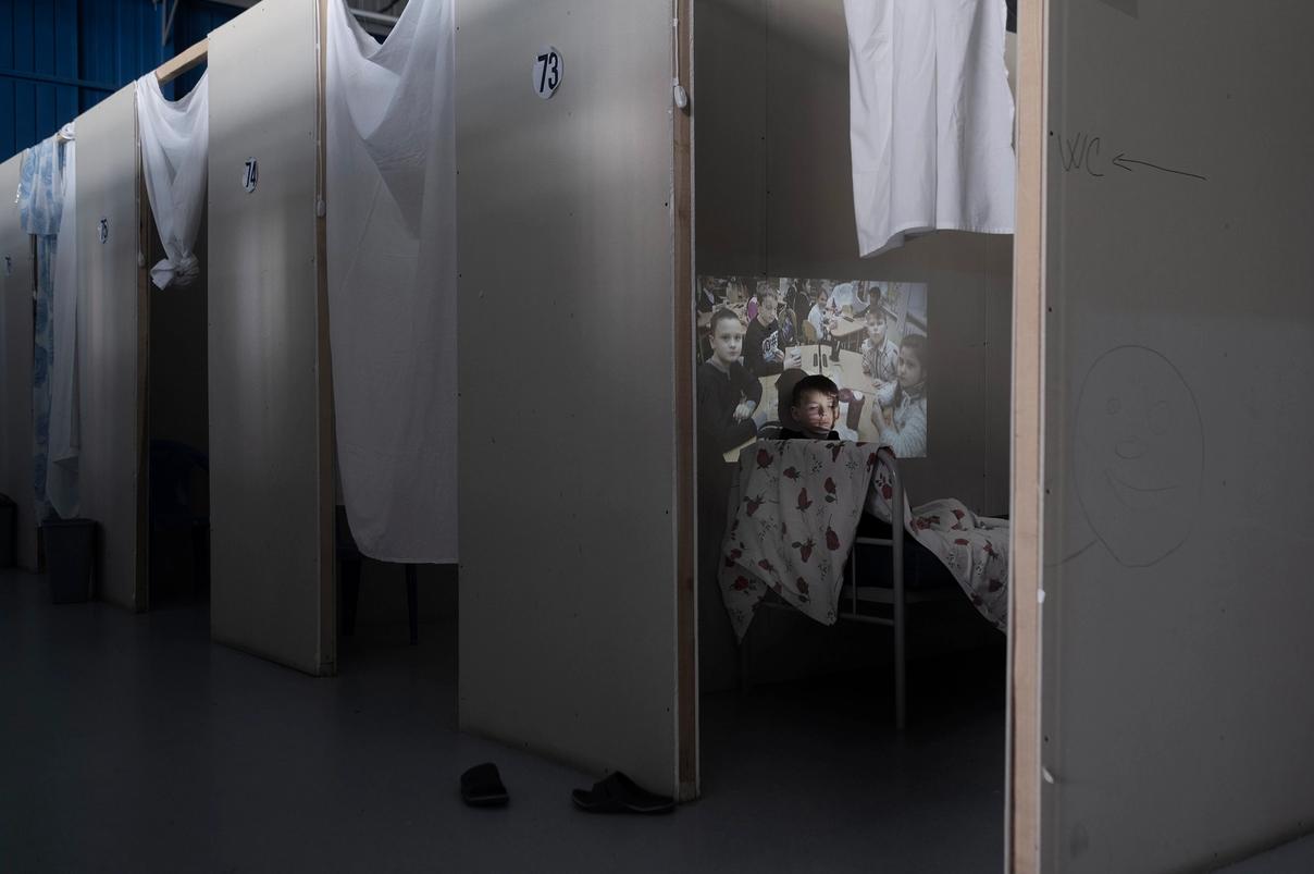 Мальчик сидит на кровати в лагере беженцев, на его лицо и стену проецируется изображение школьного класса (Poika istuu vuoteella pakolaisleirillä, hänen kasvoilleen ja seinälle heijastuu kuva koululuokasta)