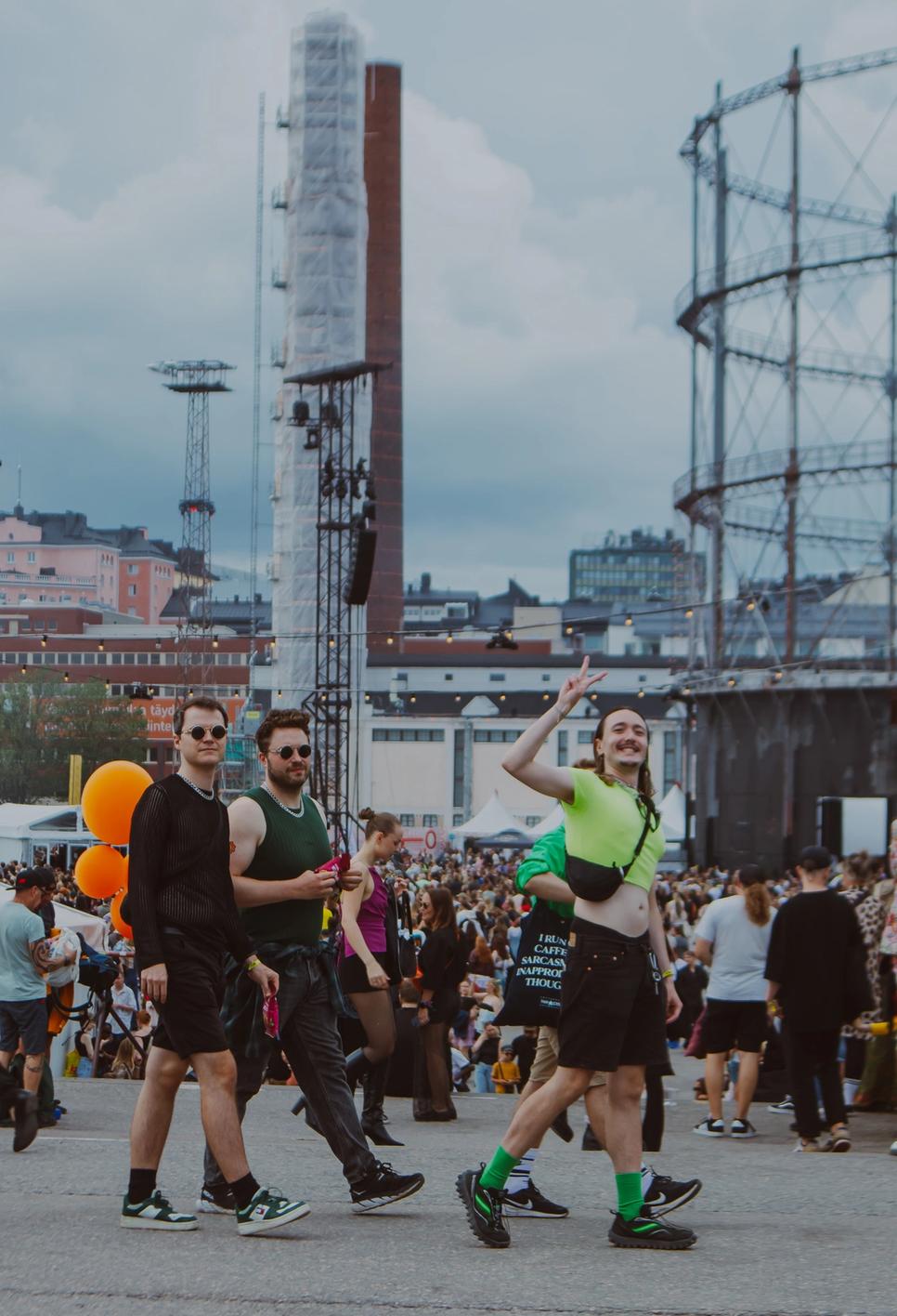 Трое молодых людей идут по фестивалю, один показывает V-жест (Kolme nuorta kävelee festivaalilla, yksi näyttää V-merkin).