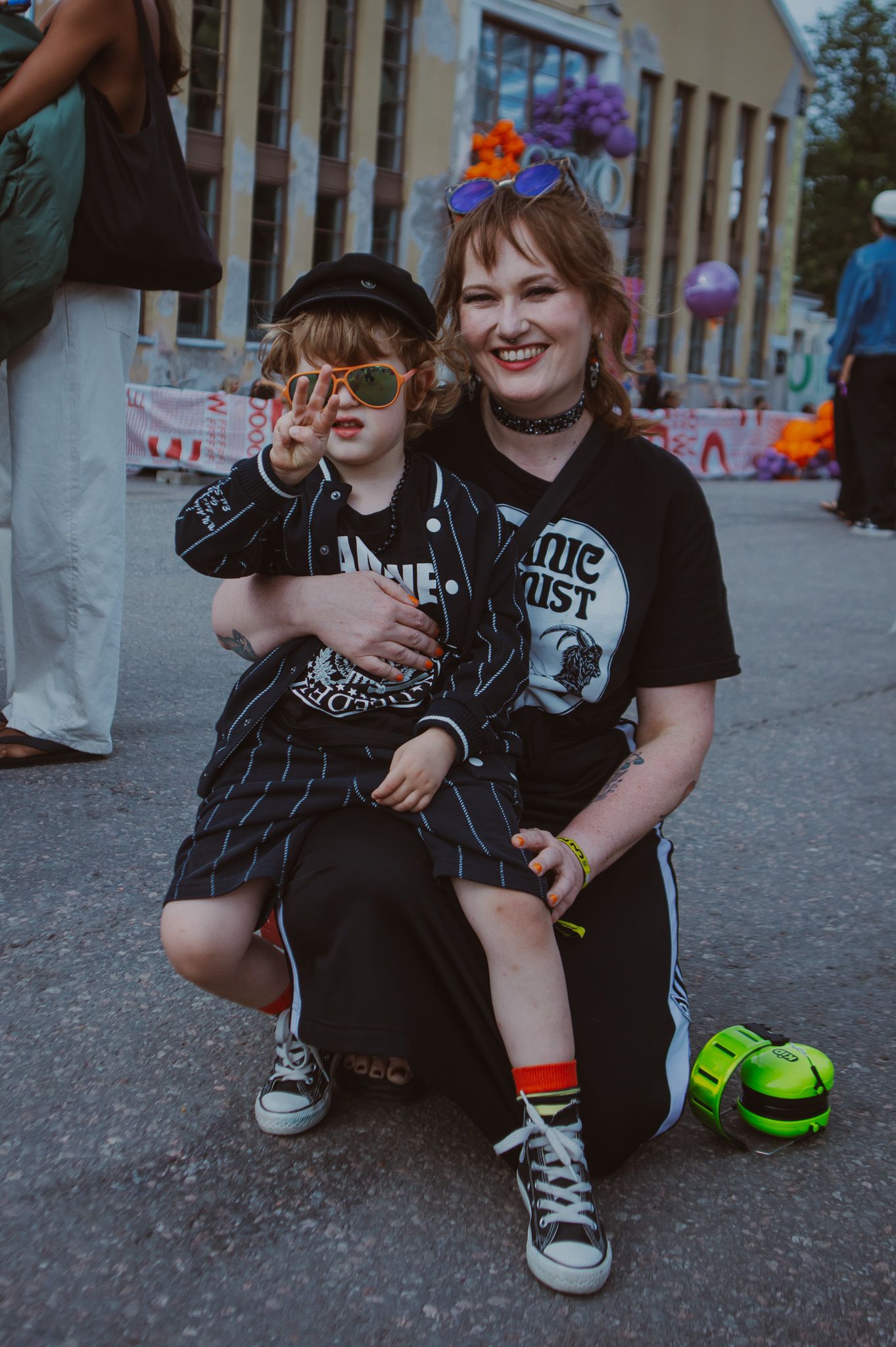 Женщина с ребёнком на фестивале, оба в тёмной одежде (Nainen ja lapsi festivaalilla tummissa vaatteissa).