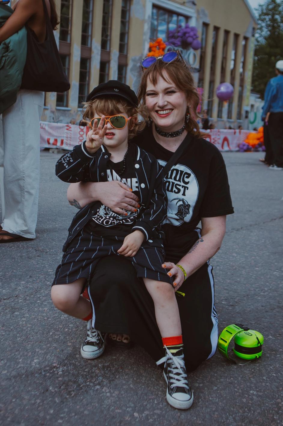 Женщина с ребёнком на фестивале, оба в тёмной одежде (Nainen ja lapsi festivaalilla tummissa vaatteissa).