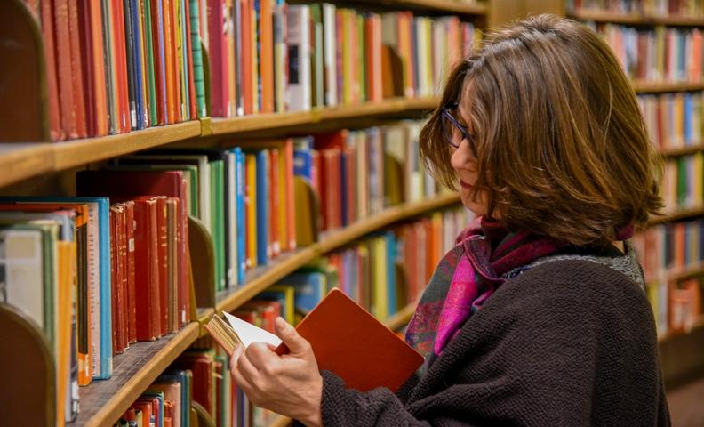 Женщина стоит в библиотеке рядом с книжной полкой и читает книгу (Nainen seisoo kirjastossa kirjahyllyn vieressä lukemassa kirjaa)
