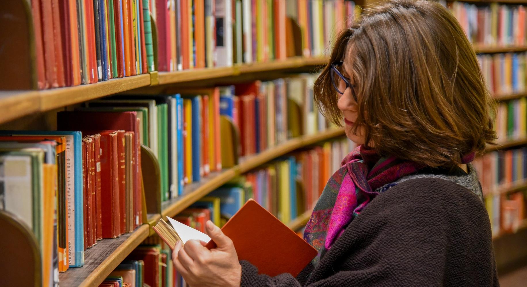 Женщина стоит в библиотеке рядом с книжной полкой и читает книгу (Nainen seisoo kirjastossa kirjahyllyn vieressä lukemassa kirjaa)