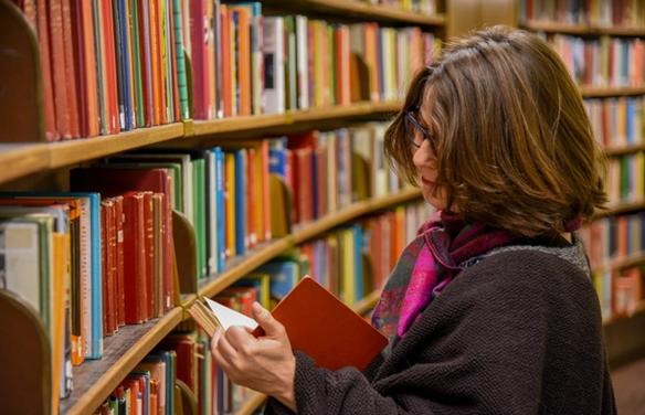 Женщина стоит в библиотеке рядом с книжной полкой и читает книгу (Nainen seisoo kirjastossa kirjahyllyn vieressä lukemassa kirjaa)