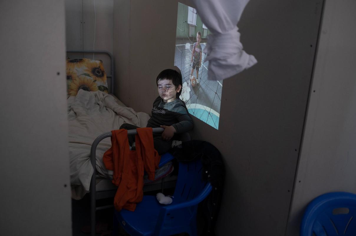 Мальчик сидит на кровати в лагере беженцев, на его лицо и стену проецируется фотография ребёнка с мячом в спортзале (Poika istuu sängyllä pakolaisleirillä, hänen kasvoilleen ja seinälle heijastuu kuva lapsesta pallon kanssa liikuntasalissa)