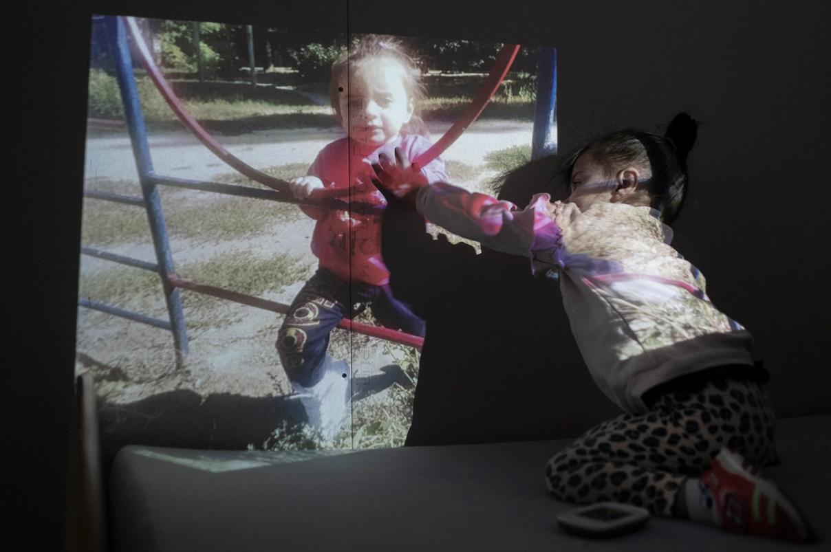 Девочка в леопардовых штанах тянется рукой к проекции фотографии, где изображён ребёнок на игровой площадке (Tyttö leopardihousuissa kurottaa kättään valokuvan heijastukseen, jossa lapsi on leikkikentällä)