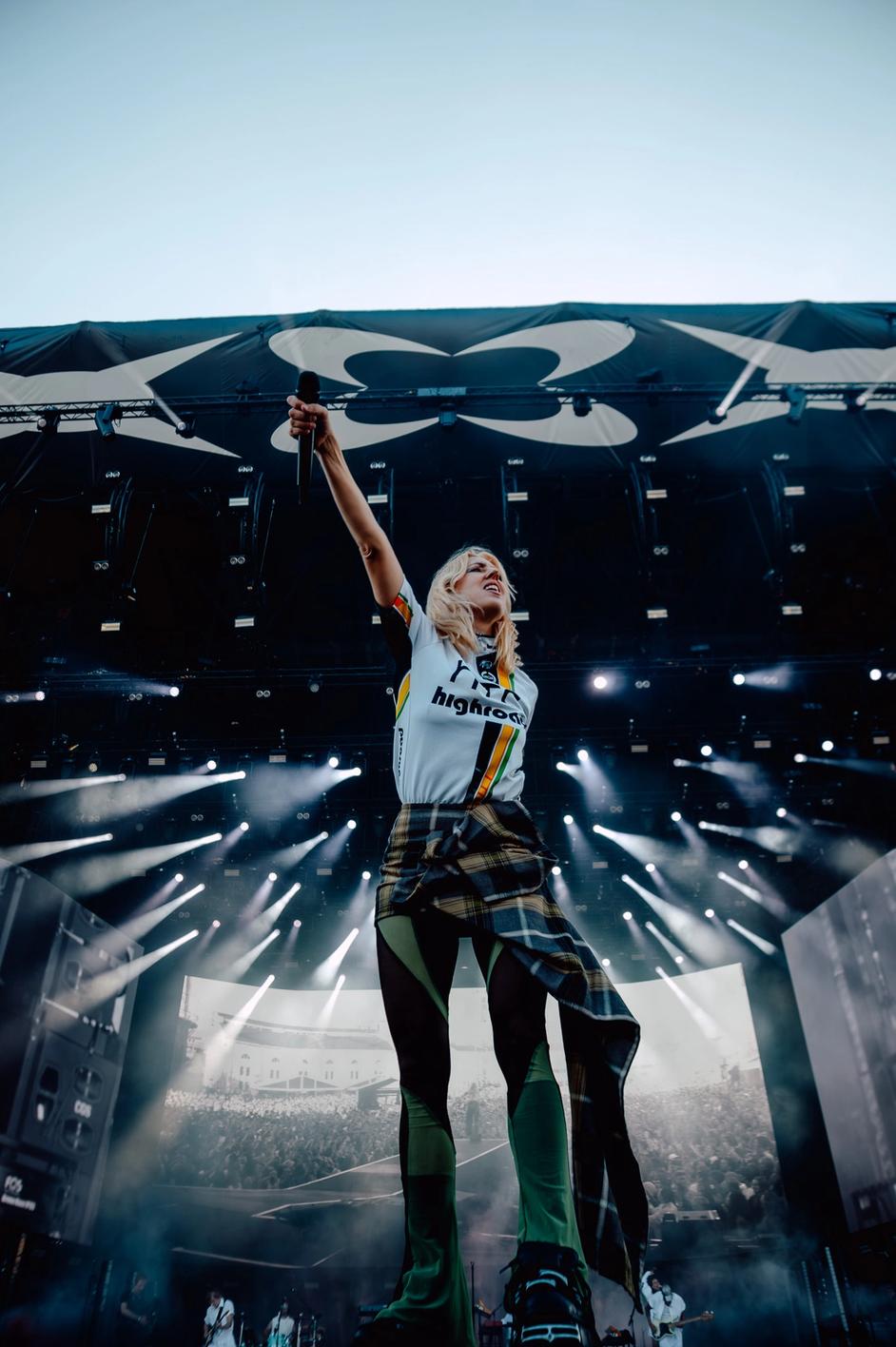 Певица на большой сцене с поднятой рукой и микрофоном среди света софитов (Laulaja suurella lavalla käsi ylhäällä valojen keskellä).